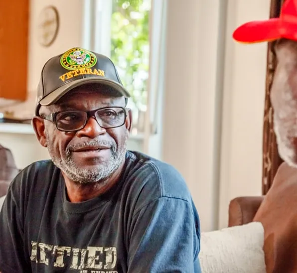 Older man wearing a veteran cap talking with another person indoors