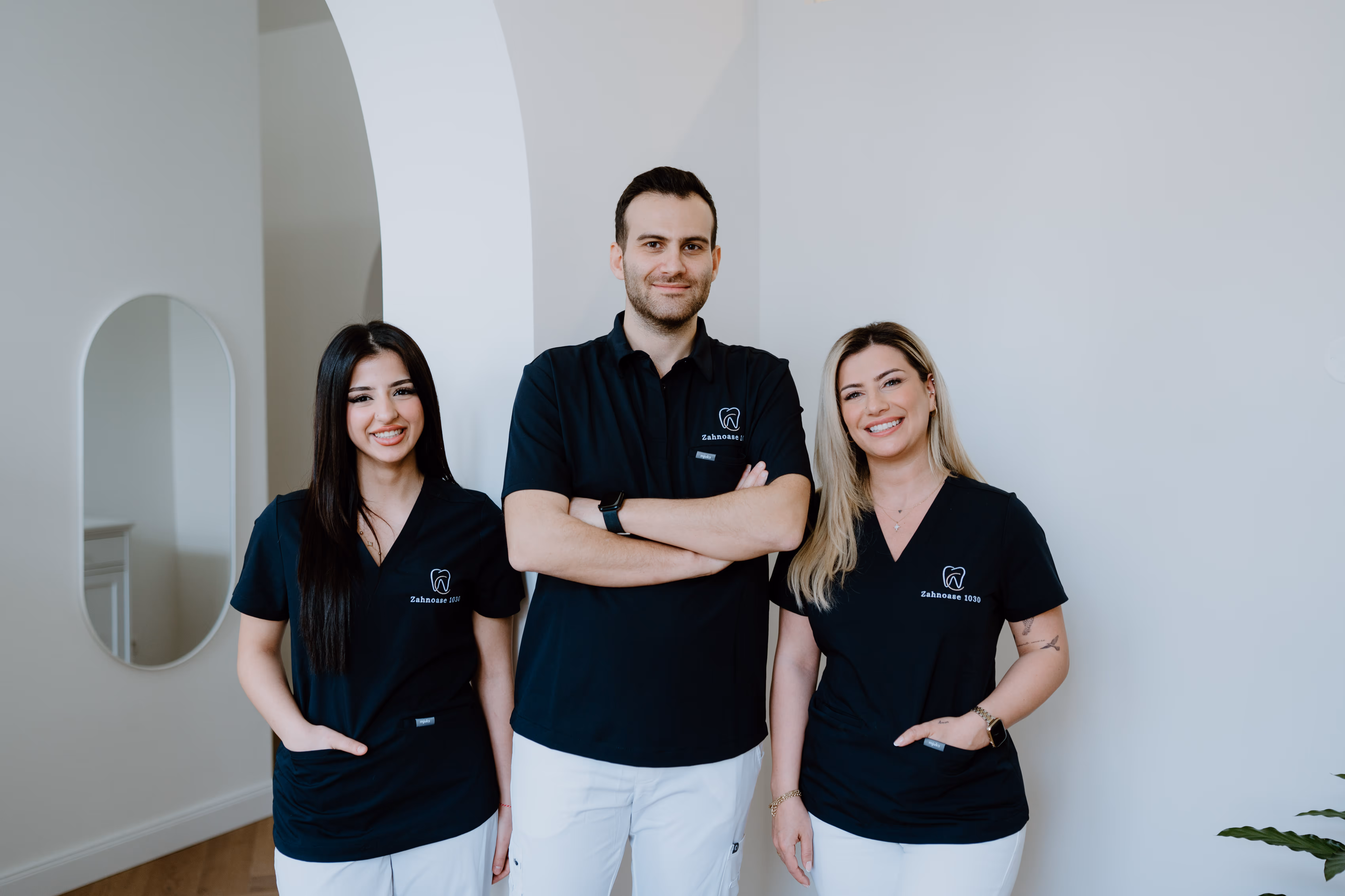Drei lächelnde Zahnärzte tragen schwarze Oberteile mit Logo und weiße Hosen vor einer weißen Wand.