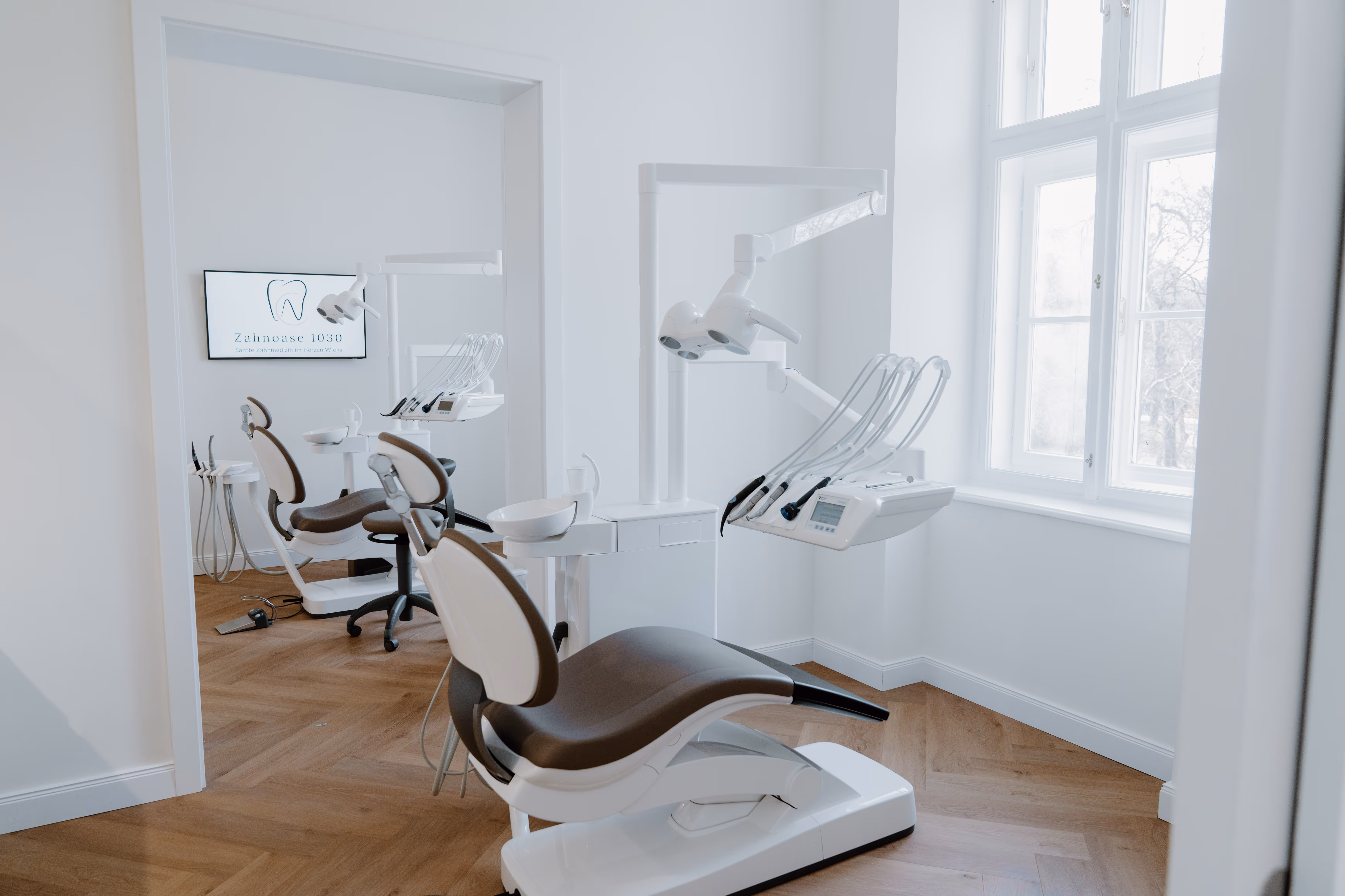 Heller, moderner Zahnarztbehandlungsraum mit zwei braunen Behandlungsstühlen, zahnärztlichen Instrumenten und Holzfußboden.