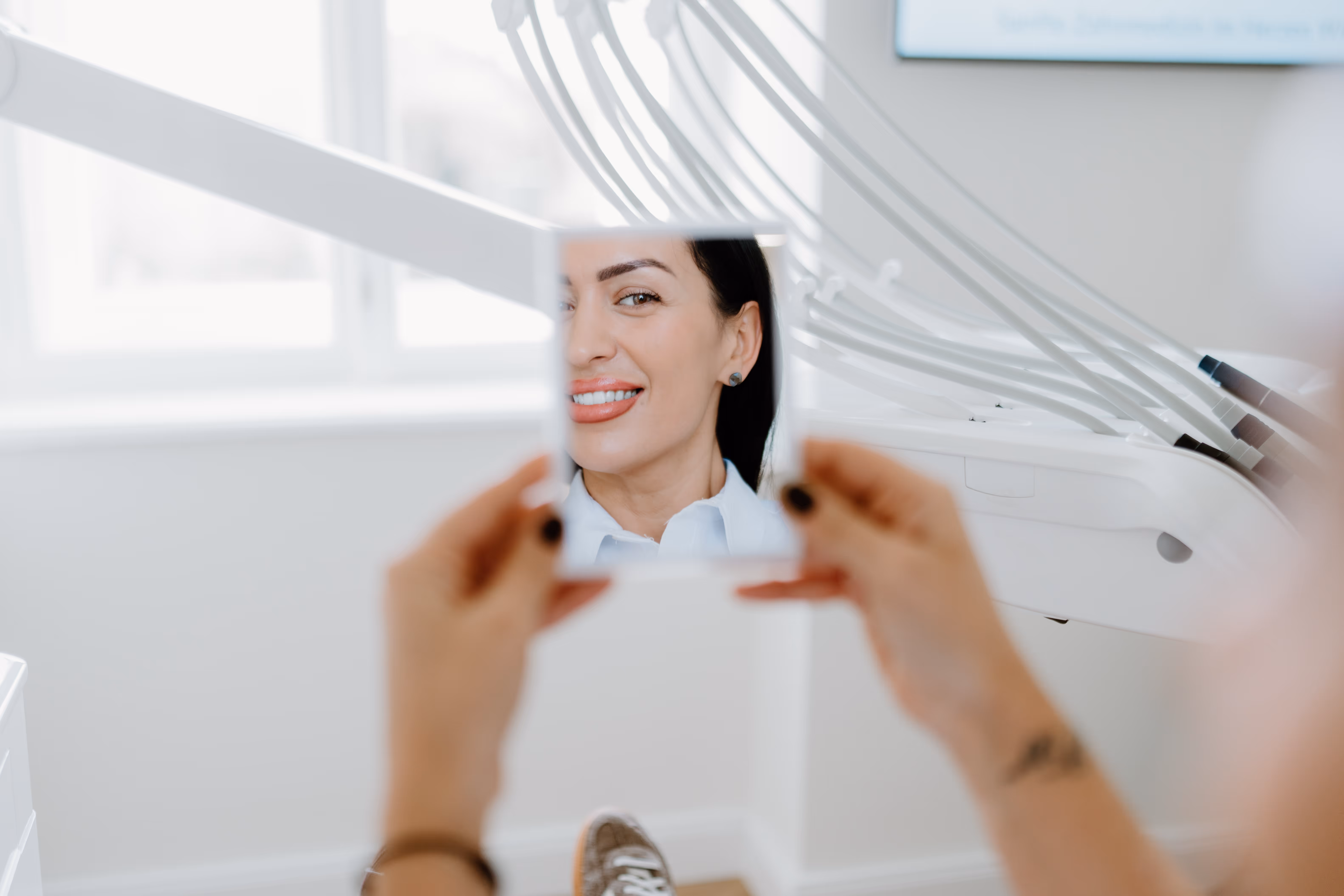 Frau lächelt und betrachtet ihr Lächeln in einem kleinen Spiegel im Zahnarztpraxis.
