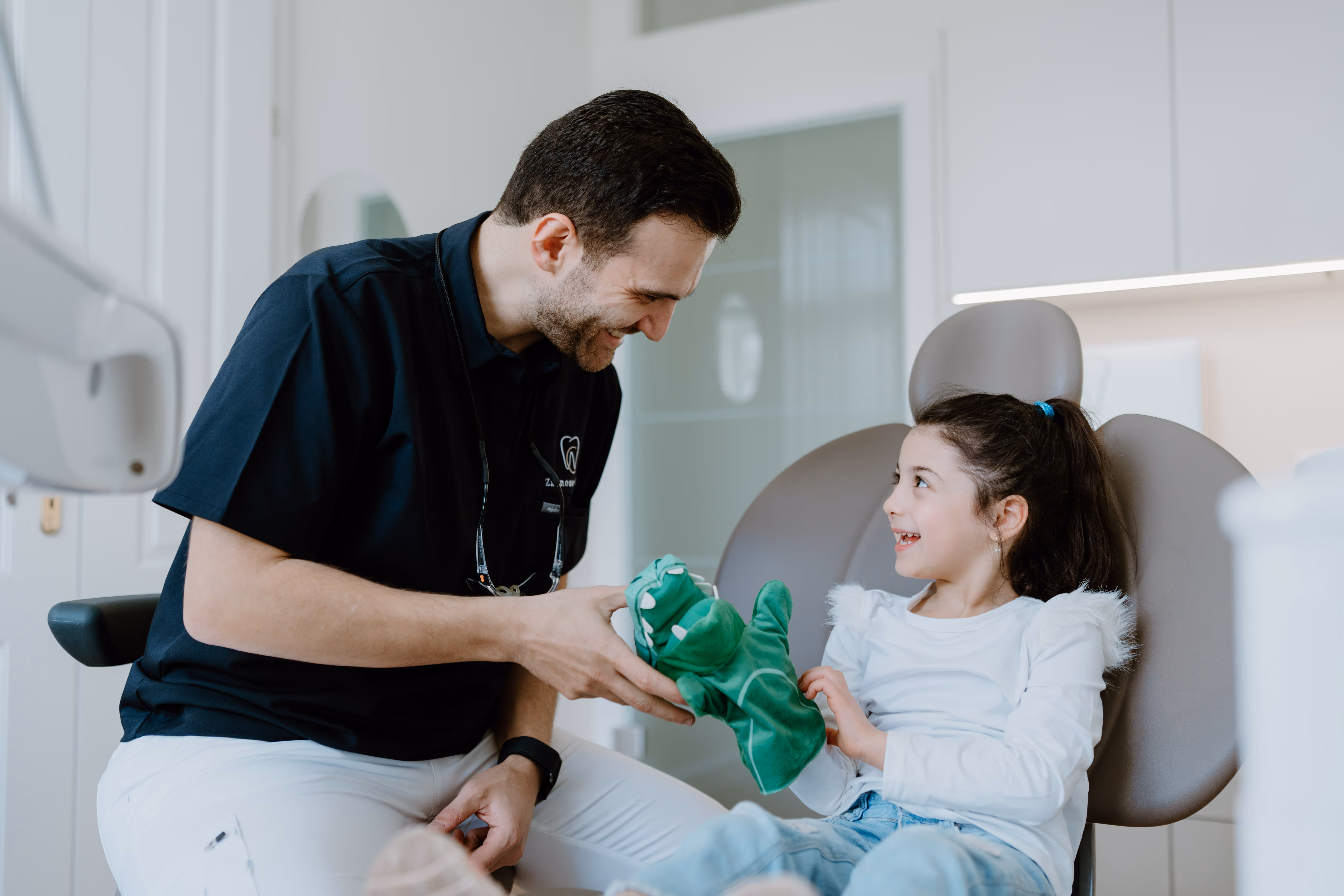 Zahnarzt unterhält sich lächelnd mit einem Mädchen in einem Behandlungsstuhl und hält eine grüne Krokodil-Handpuppe.