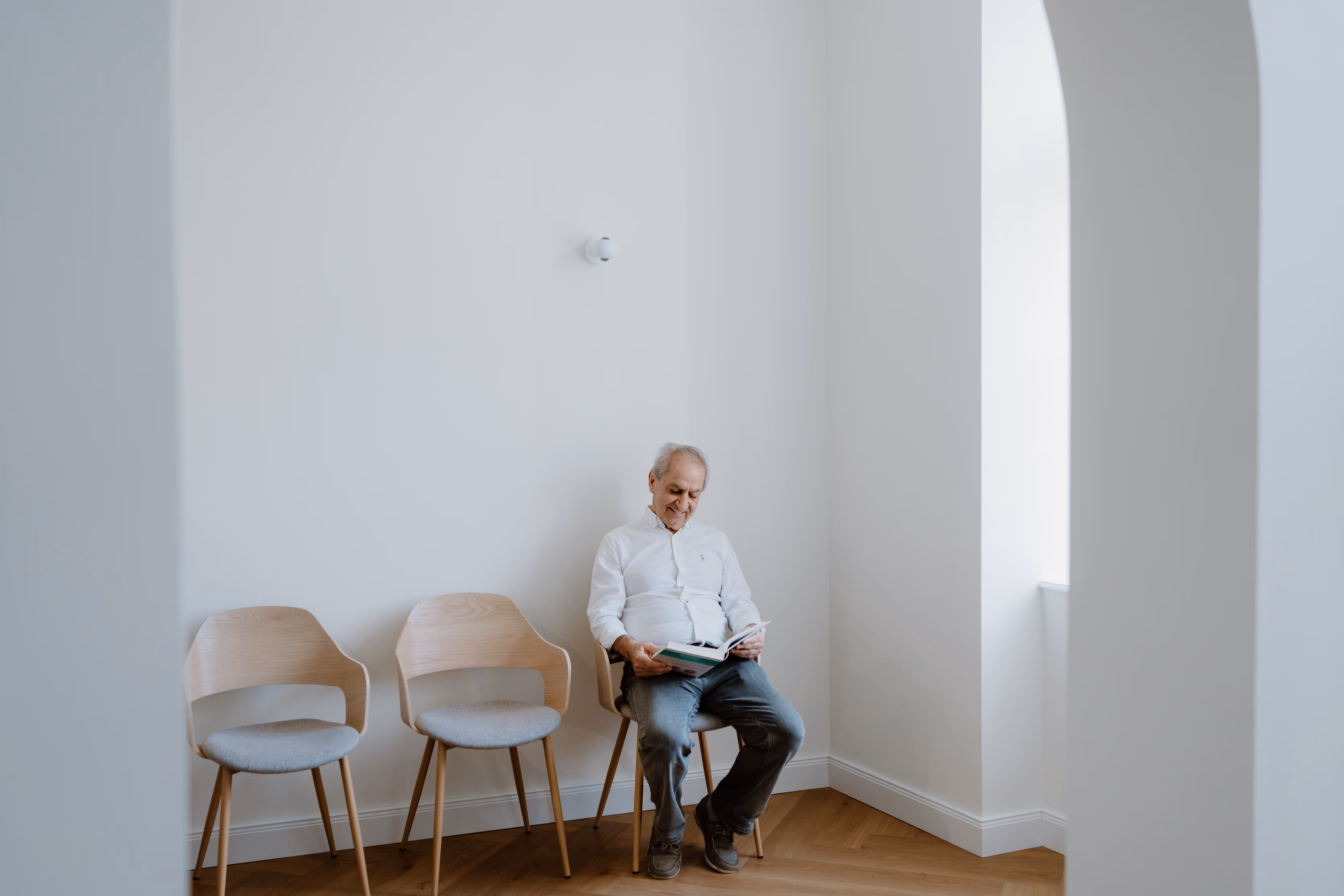 Älterer Mann sitzt auf einem Stuhl in einem hellen, minimalistischen Raum und liest ein Buch.