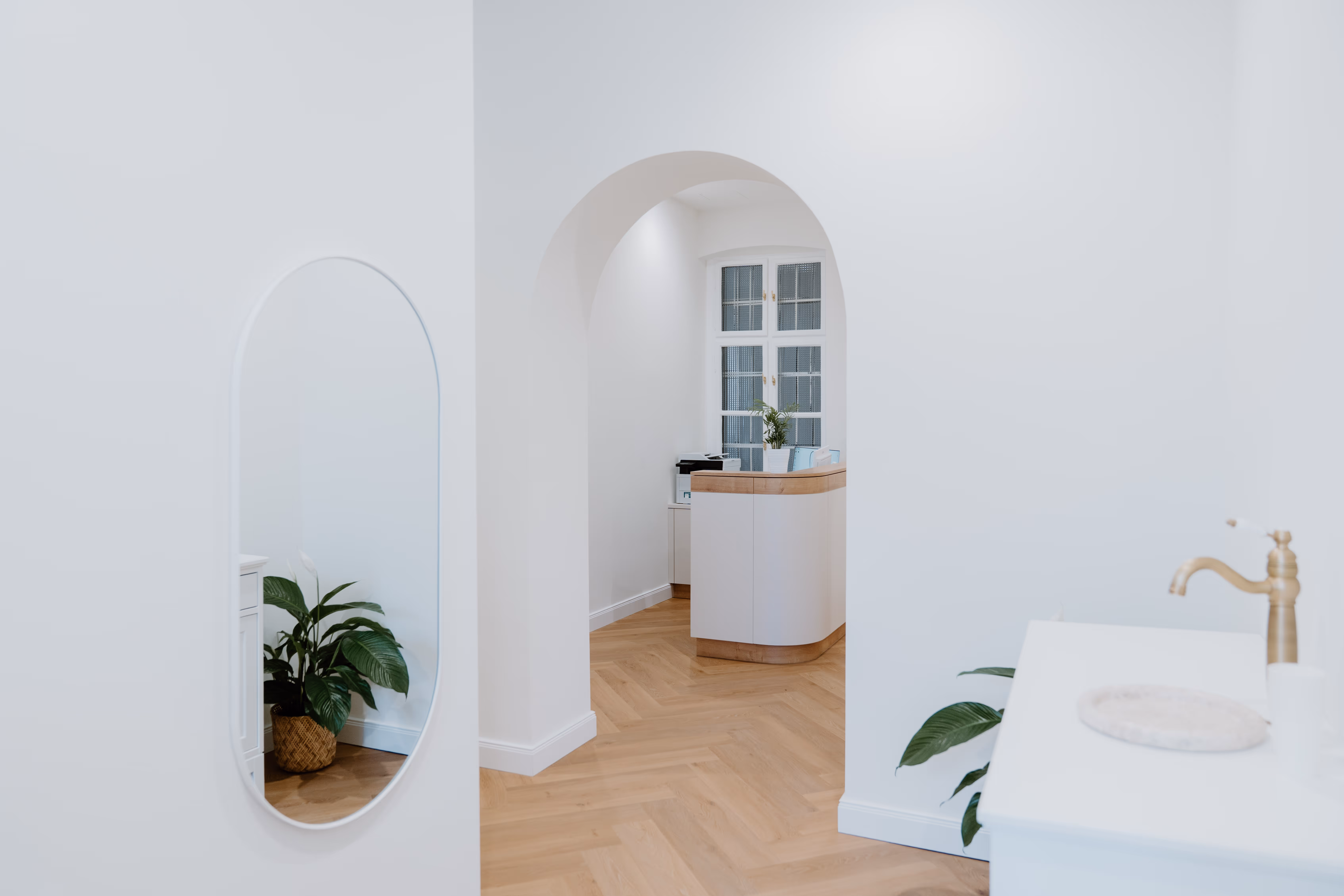 Heller Raum mit weiß gestrichenen Wänden, Holzboden, ovalem Wandspiegel, Pflanzen und Empfangstheke unter einem Bogen.