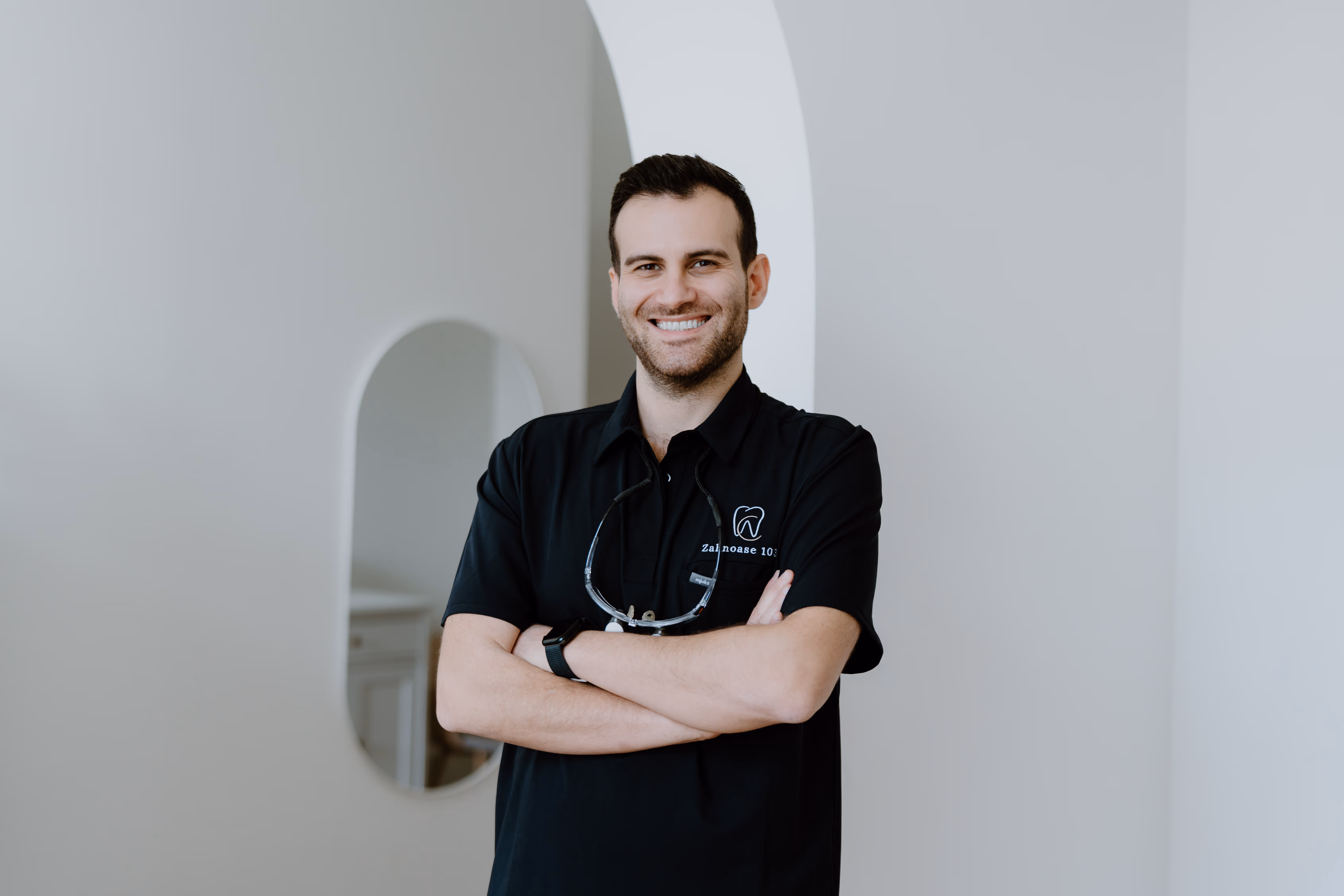 Lächelnder Zahnarzt mit verschränkten Armen in schwarzem Poloshirt mit Zahnarzt-Logo vor hellem Hintergrund.