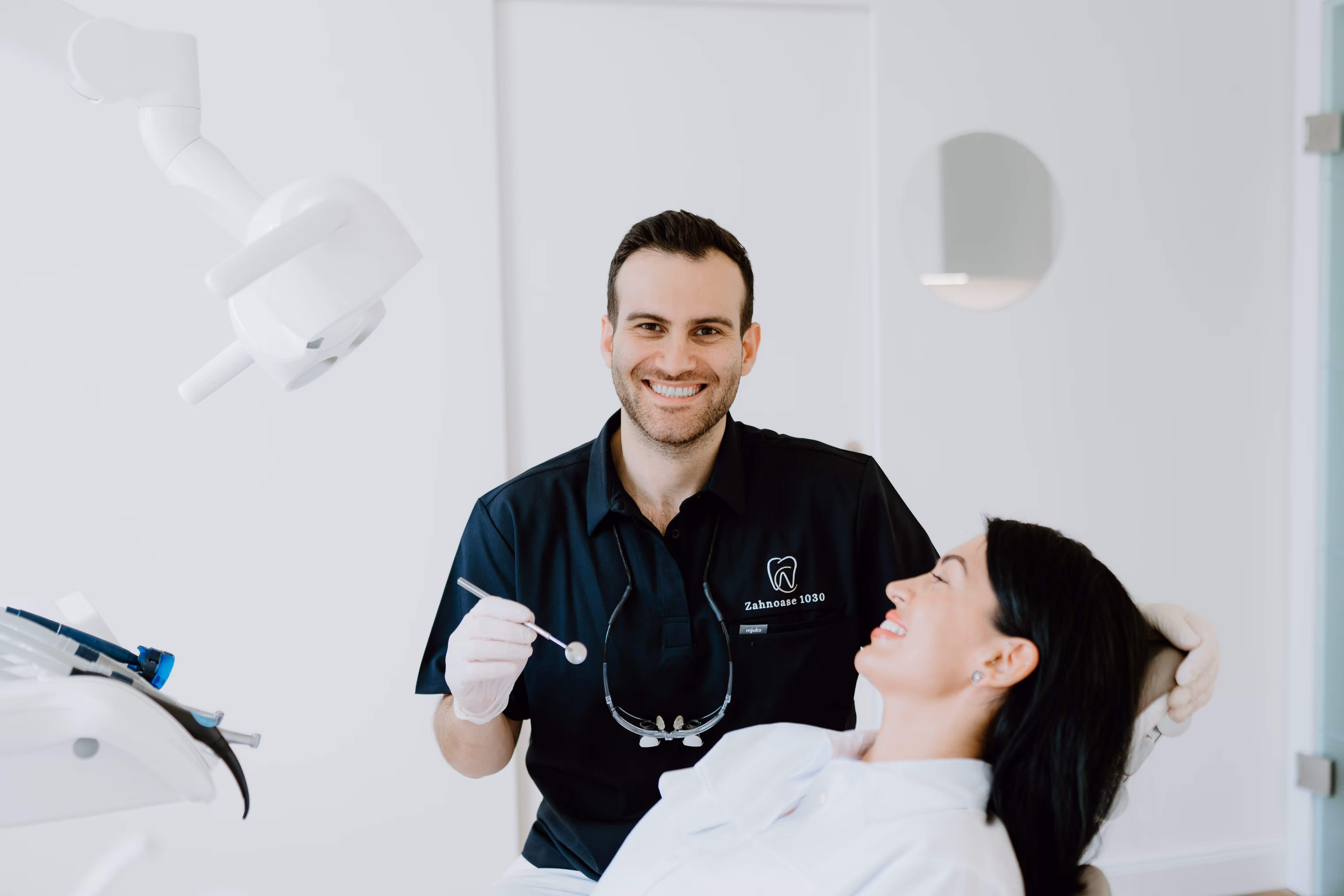 Zahnarzt im schwarzen Kittel lächelt, während Patientin mit dunklen Haaren auf dem Behandlungsstuhl sitzt und ebenfalls lächelt.