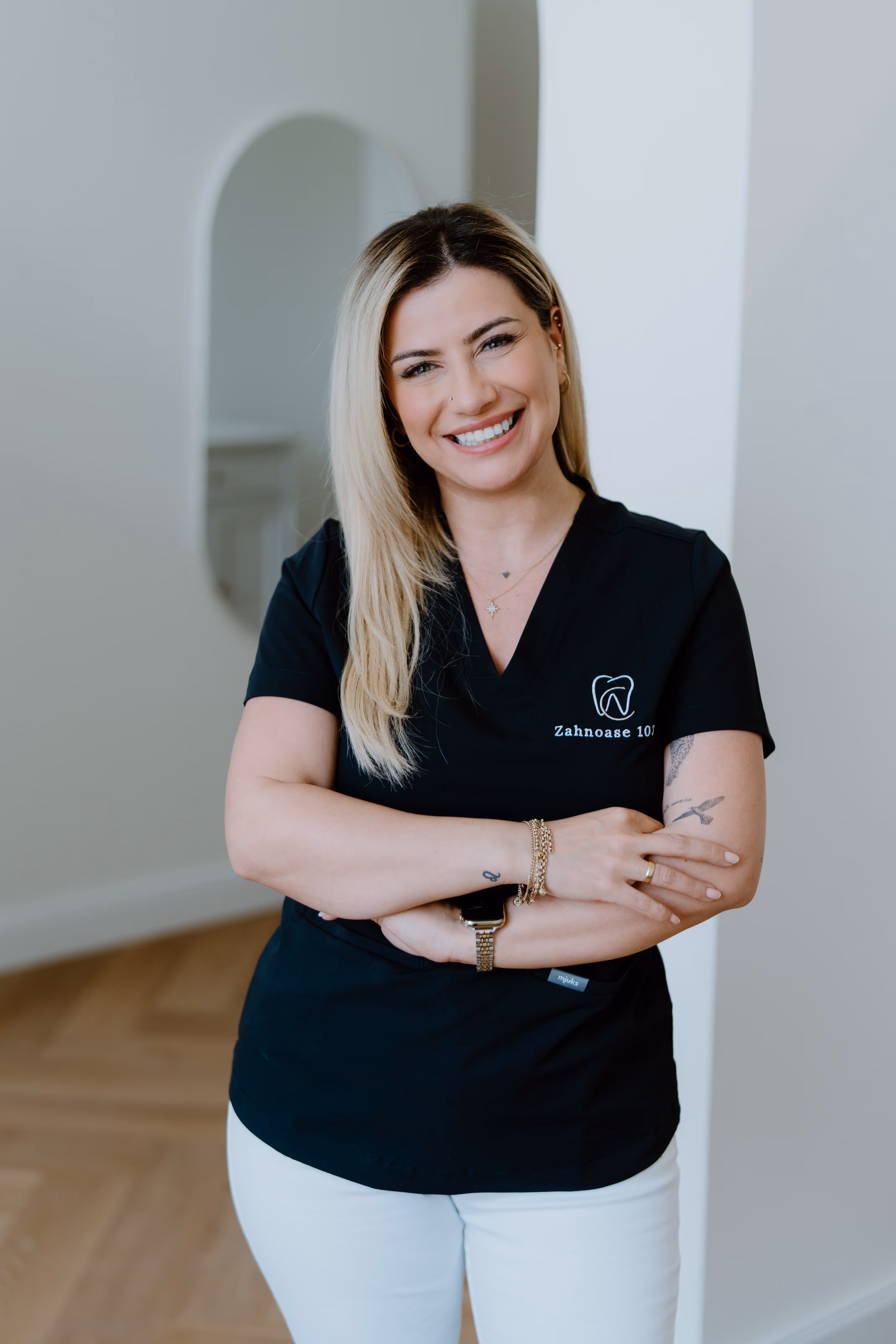 Fröhliche Zahnärztin mit blondem Haar trägt schwarze Arbeitskleidung mit Zahnarztlogo und weiße Hose, steht mit verschränkten Armen.