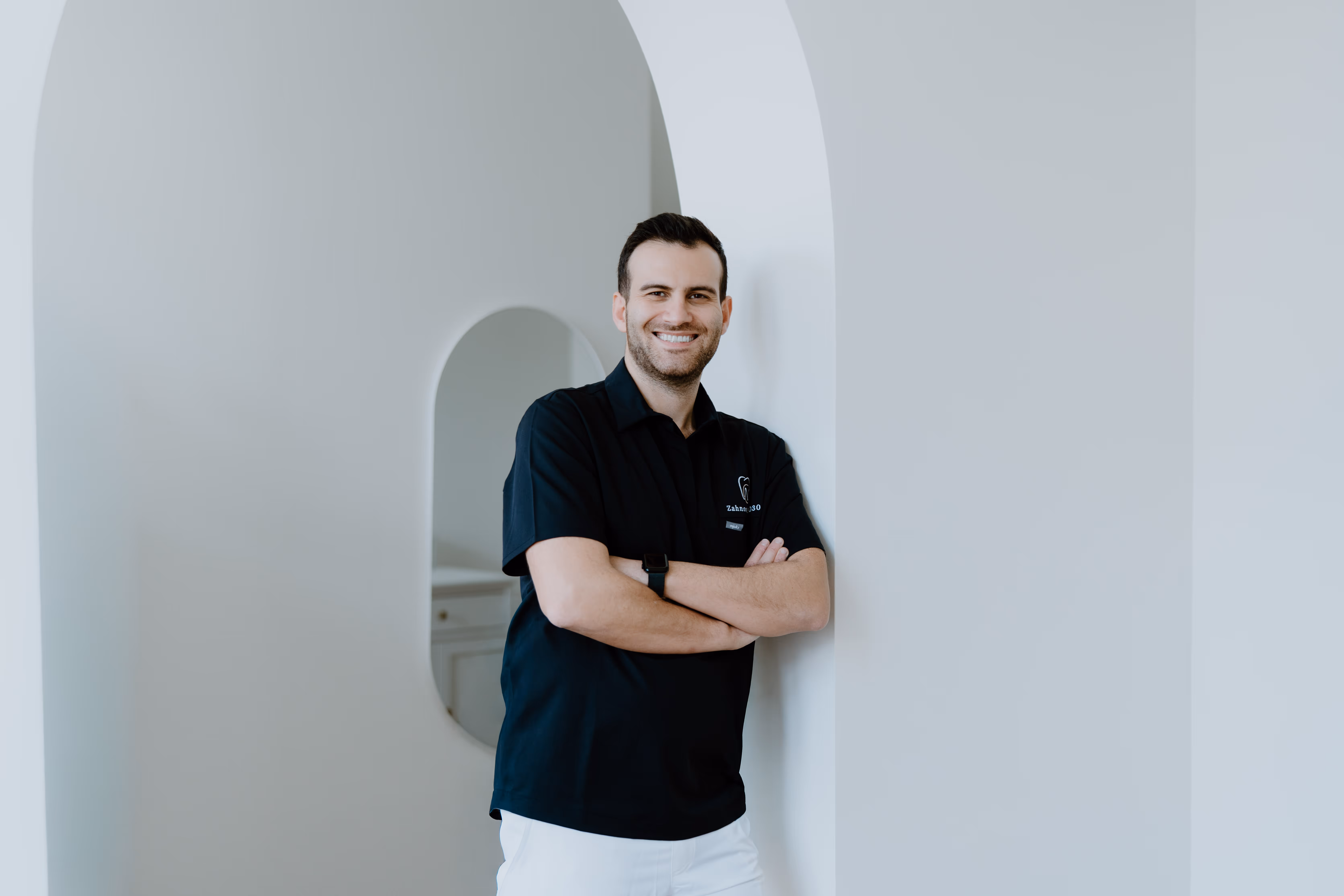 Lächelnder Mann in schwarzem Polohemd steht mit verschränkten Armen vor weißer Wand mit Bogen.