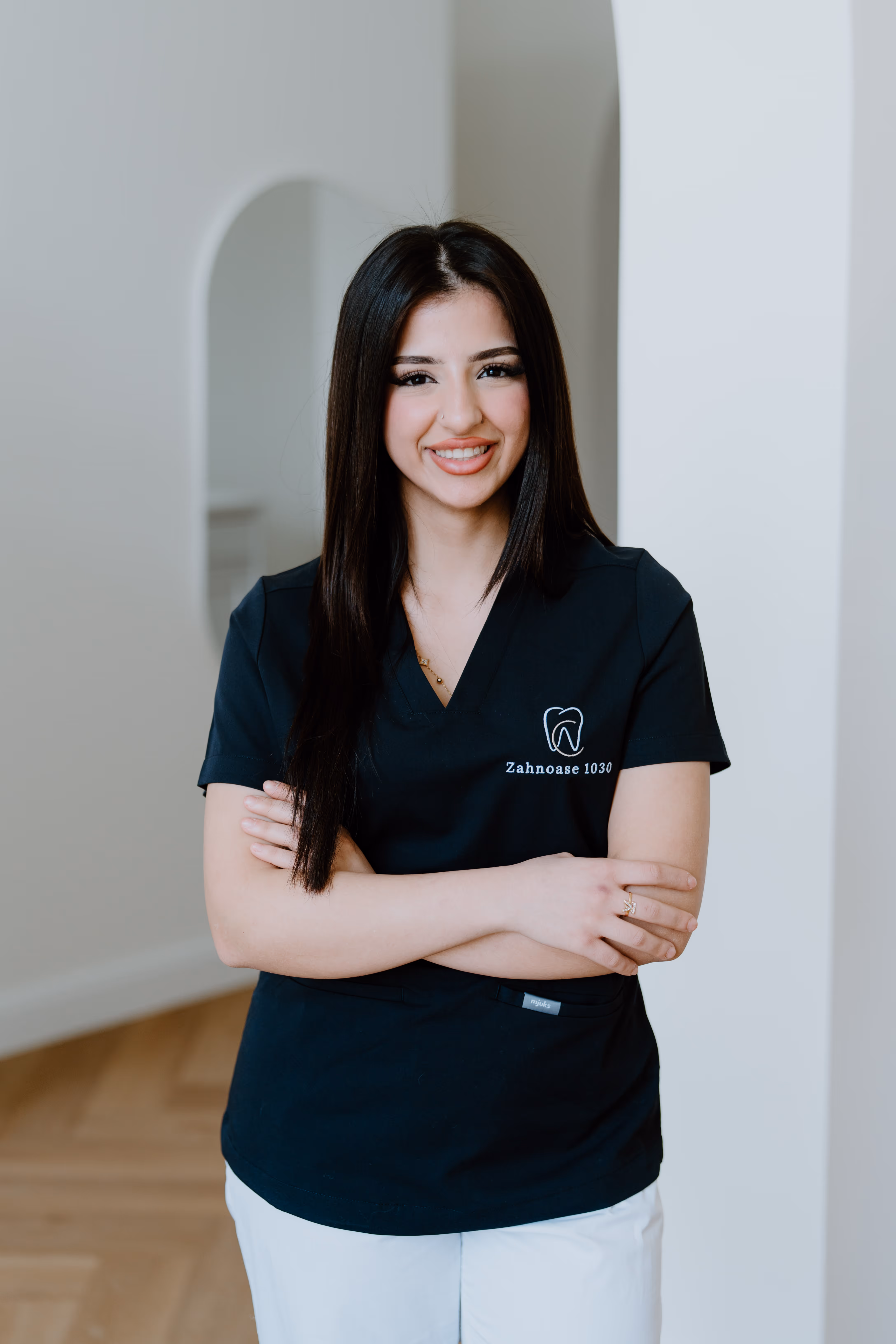 Junge Zahnärztin mit langen dunklen Haaren in schwarzer Arbeitskleidung mit Logo von Zahnoase 1030, lächelnd mit verschränkten Armen.