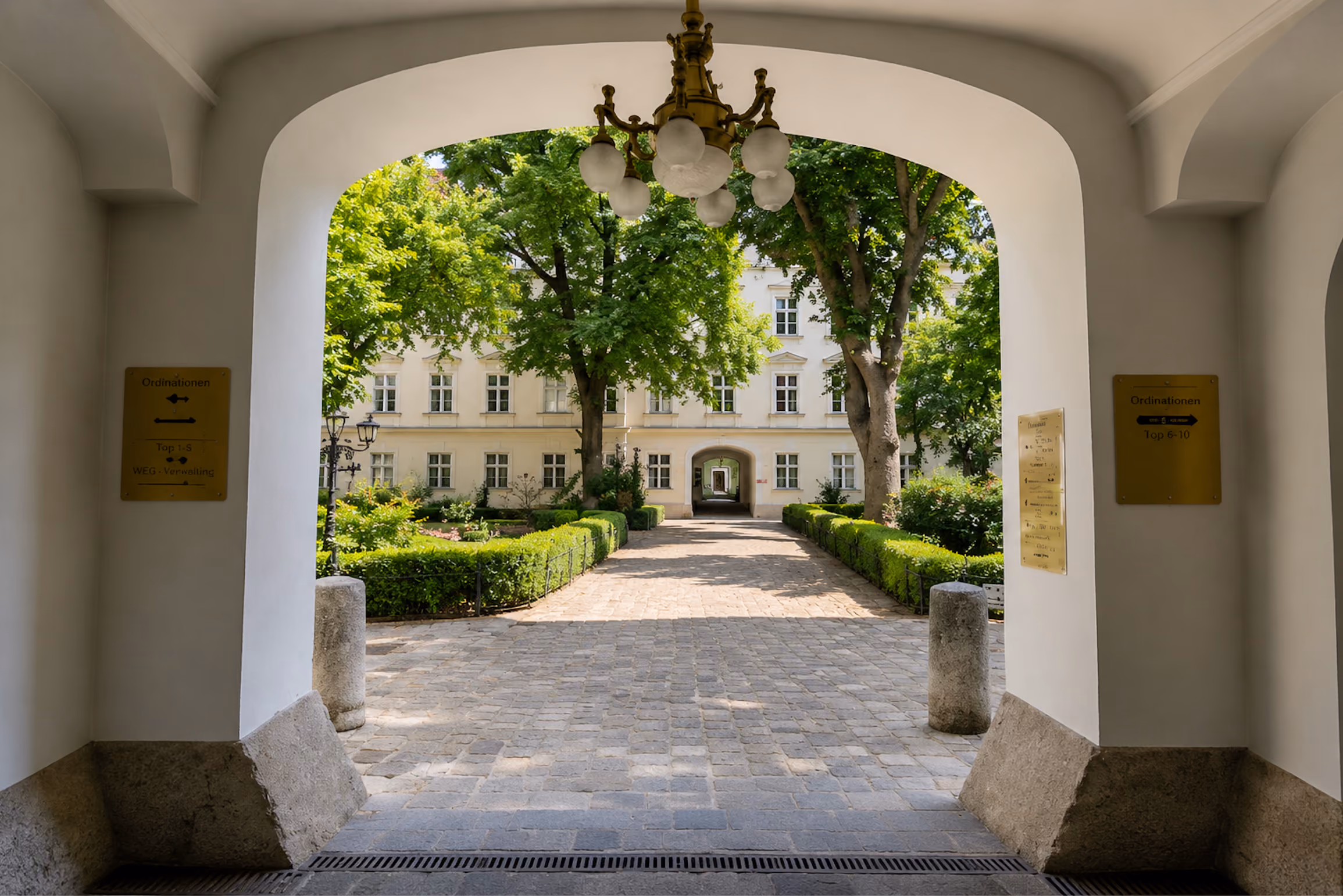 Blick durch einen Torbogen auf gepflasterten Weg, grüne Hecken, Bäume und ein weißes Gebäude mit Fenstern.