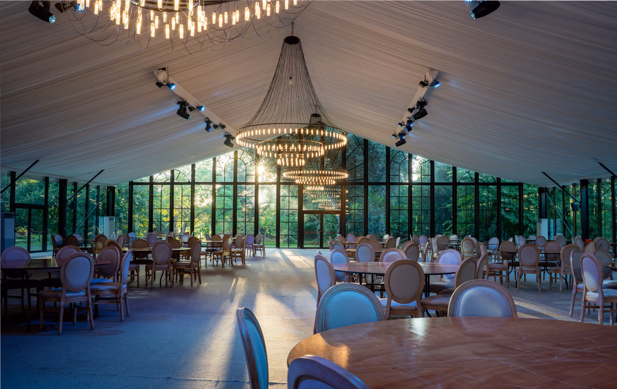 Spacious event hall with round tables and chairs under illuminated chandeliers and large glass windows showing green trees outside.