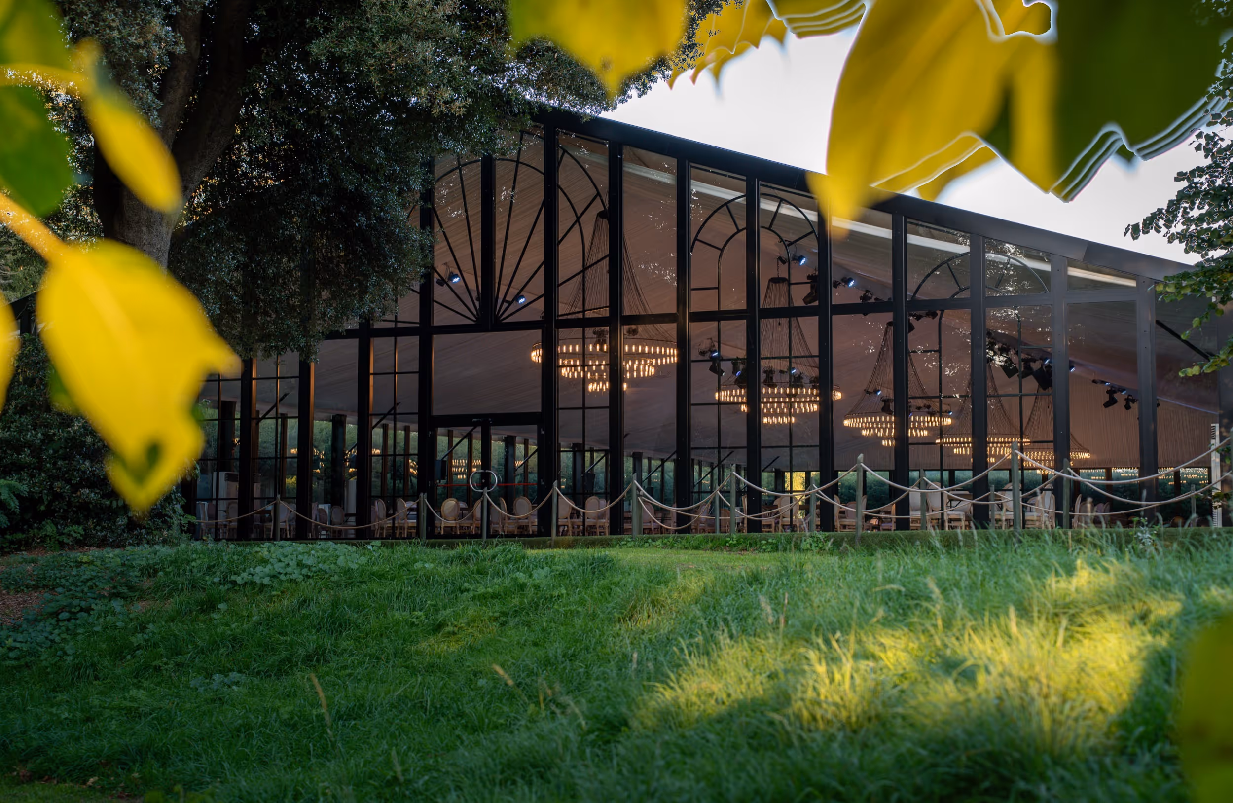 Glass-walled event tent with chandeliers inside, surrounded by green grass and trees.