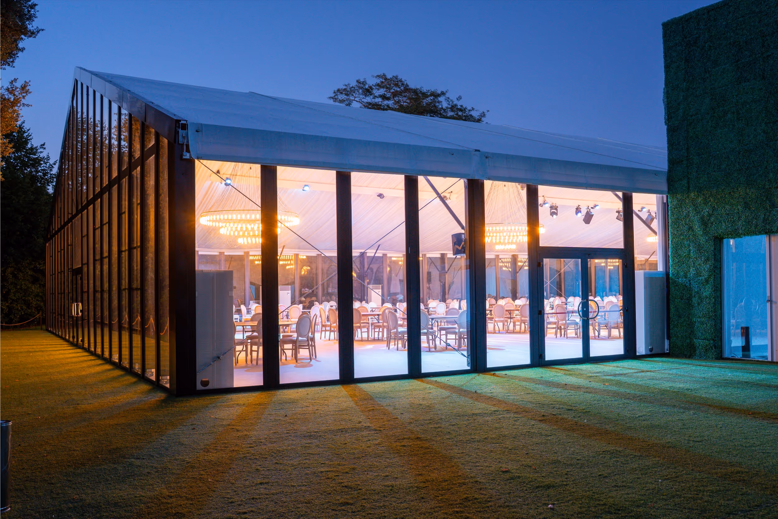 Large glass-walled event tent illuminated at dusk with round dining tables and chairs inside.