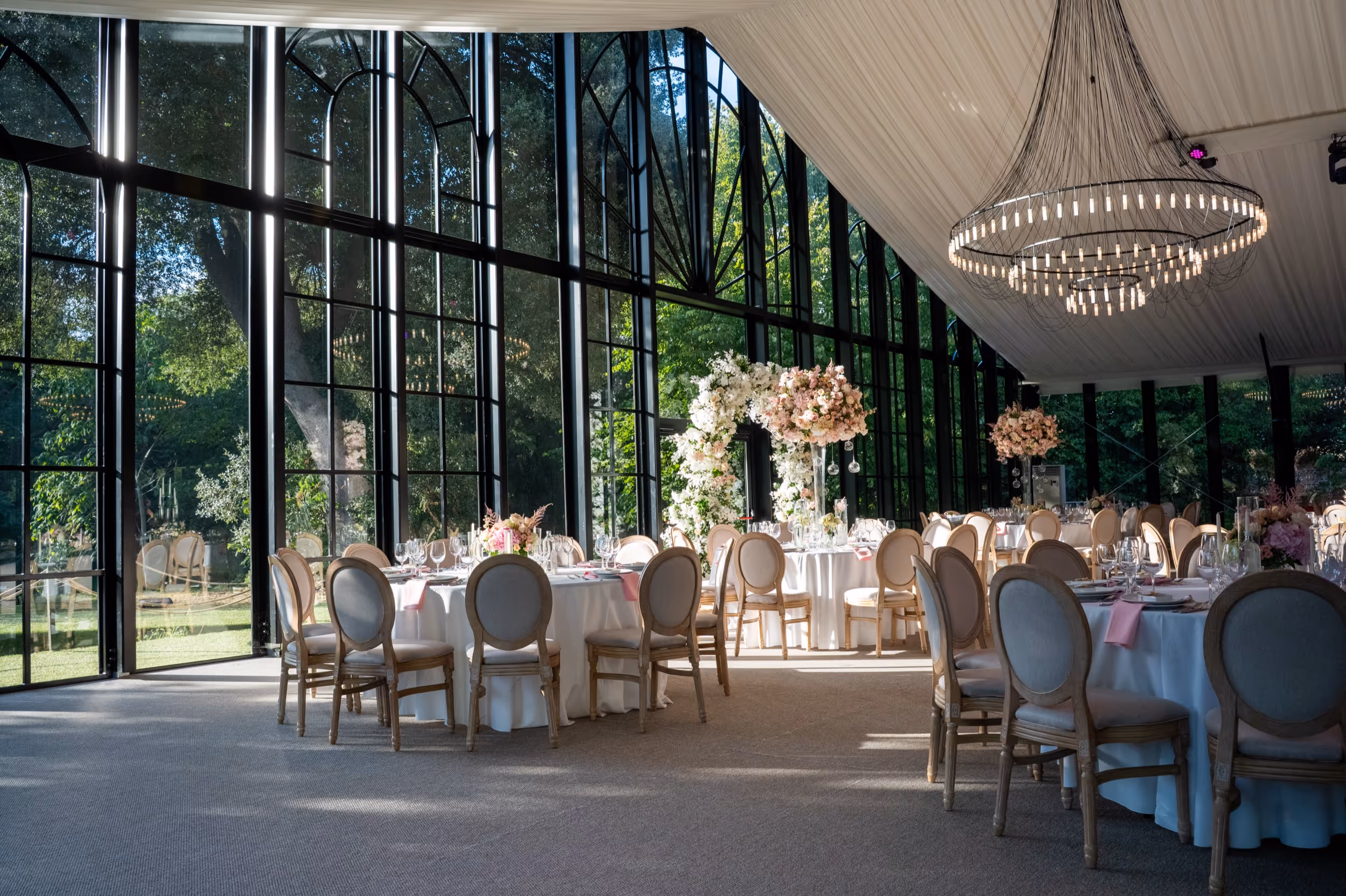 Elegant wedding reception setup with round tables, wooden chairs, large windows showing greenery, floral arrangements, and a modern chandelier.