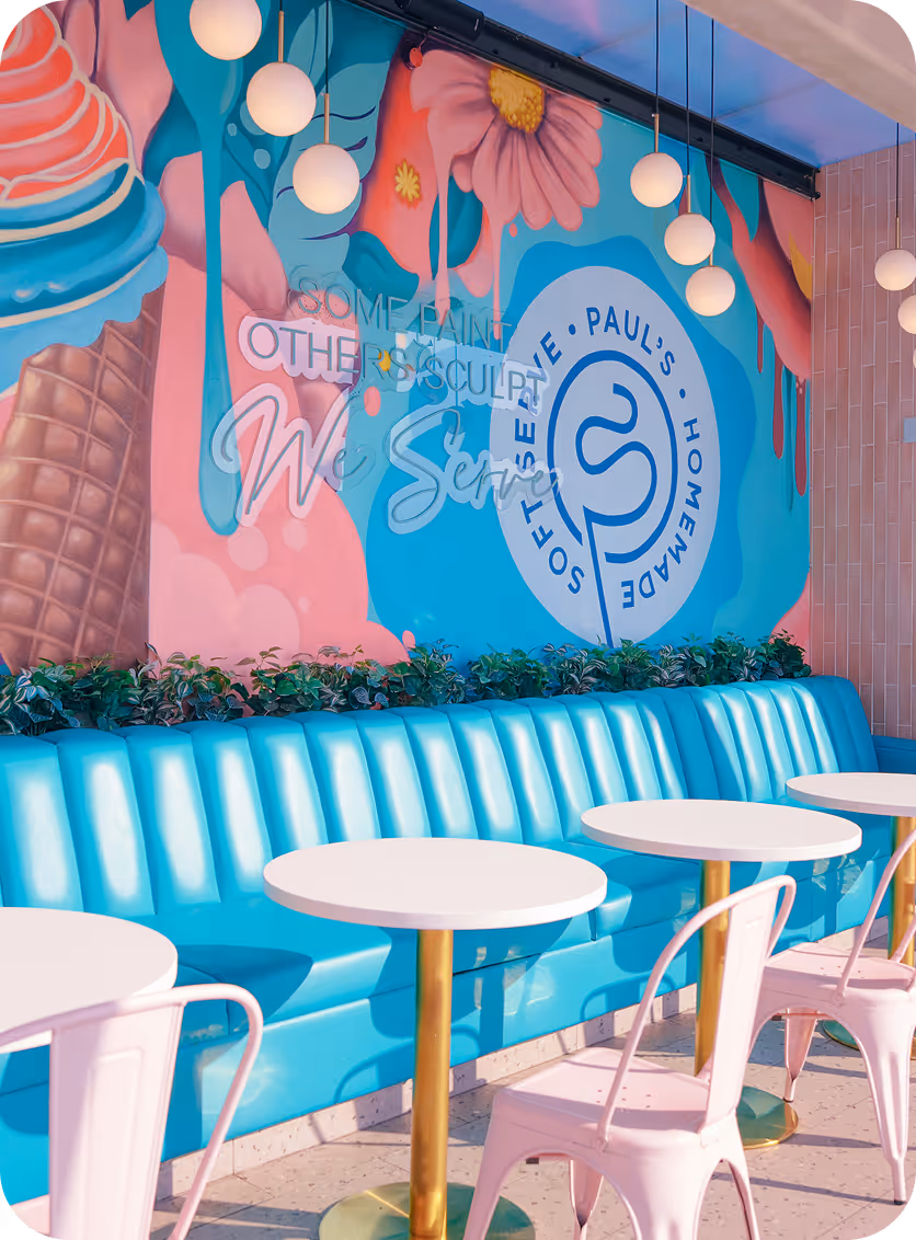 Bright cafe interior with turquoise booth seating, white round tables, pink chairs, and colorful wall mural featuring ice cream cone and flowers.