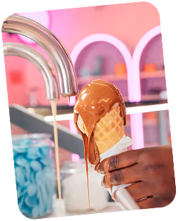 Hand holding an ice cream cone being covered with flowing melted chocolate in a shop with pink neon lighting.