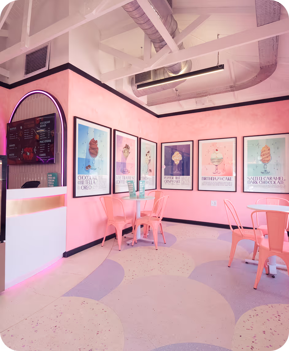 Pink-themed ice cream parlor interior with pastel pink walls, framed menu posters, and pink metal chairs around white tables.