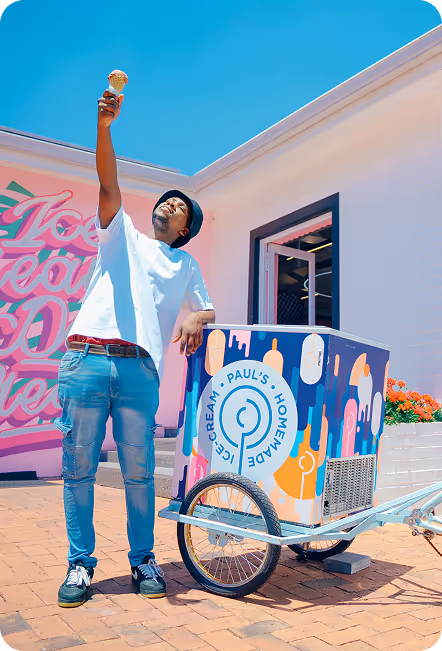 Man in white t-shirt and jeans holding up an ice cream cone next to a colorful ice cream cart on a sunny day.
