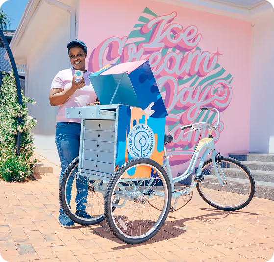 Man in white t-shirt and jeans holding an ice cream cone up beside a colorful Paul’s Homemade Ice Cream cart outdoors.
