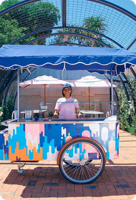 Man in a white t-shirt and jeans holding an ice cream cone up next to a colorful ice cream cart labeled 'Paul's Homemade Ice Cream' under a clear blue sky.