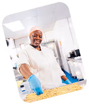 Smiling food factory worker wearing a hairnet and gloves spreading cereal on a tray in a clean industrial kitchen.