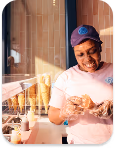 Smiling female chef in white uniform and pink headscarf spreading dough or crumbs on a tray in a professional kitchen.