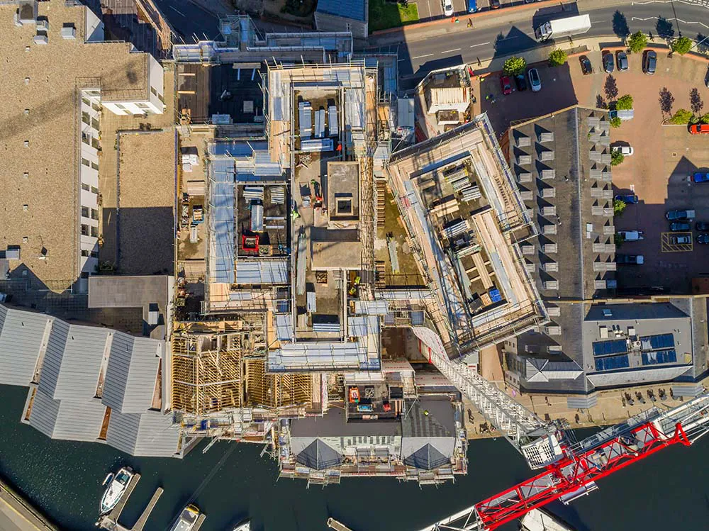 Aerial view of a large construction site next to a harbor with several buildings, cranes, parked cars, and boats in the water.