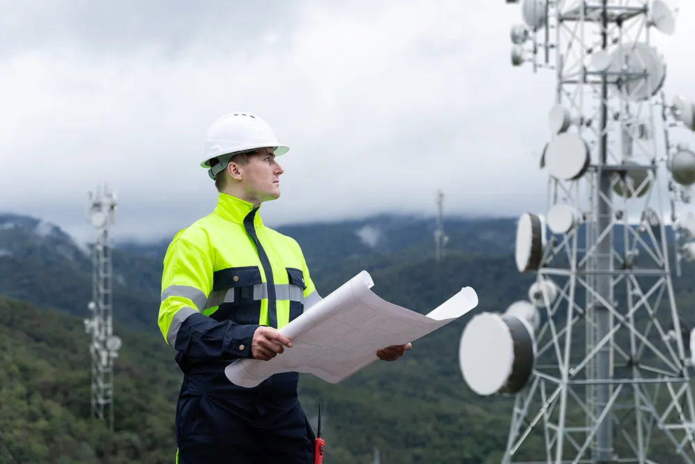 Engineer in high-visibility jacket and white helmet holding blueprints near telecom towers in a mountainous area.