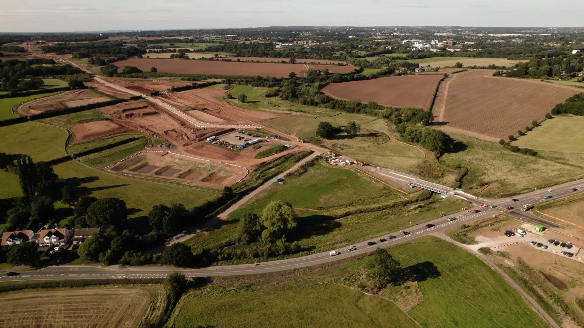 Aerial view of rural road construction site with fields, vehicles, and scattered trees, installing linear infrastructure