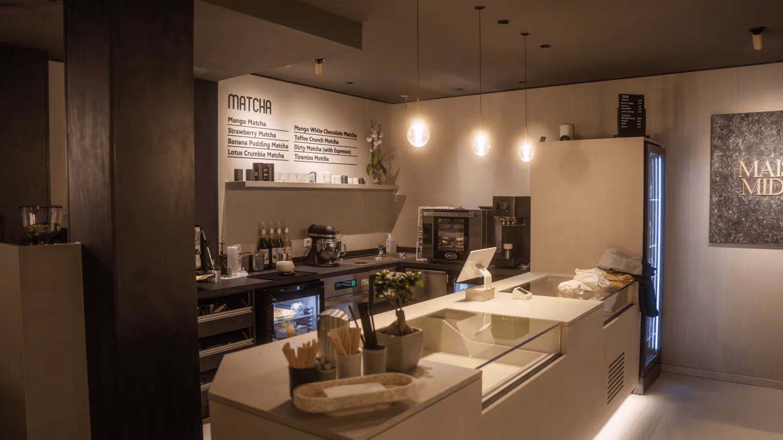 Modernes, gedämpft beleuchtetes Café mit einer Theke, Kaffeemaschine, komfortabler Ausstattung und einer Menütafel für Matcha-Getränke.