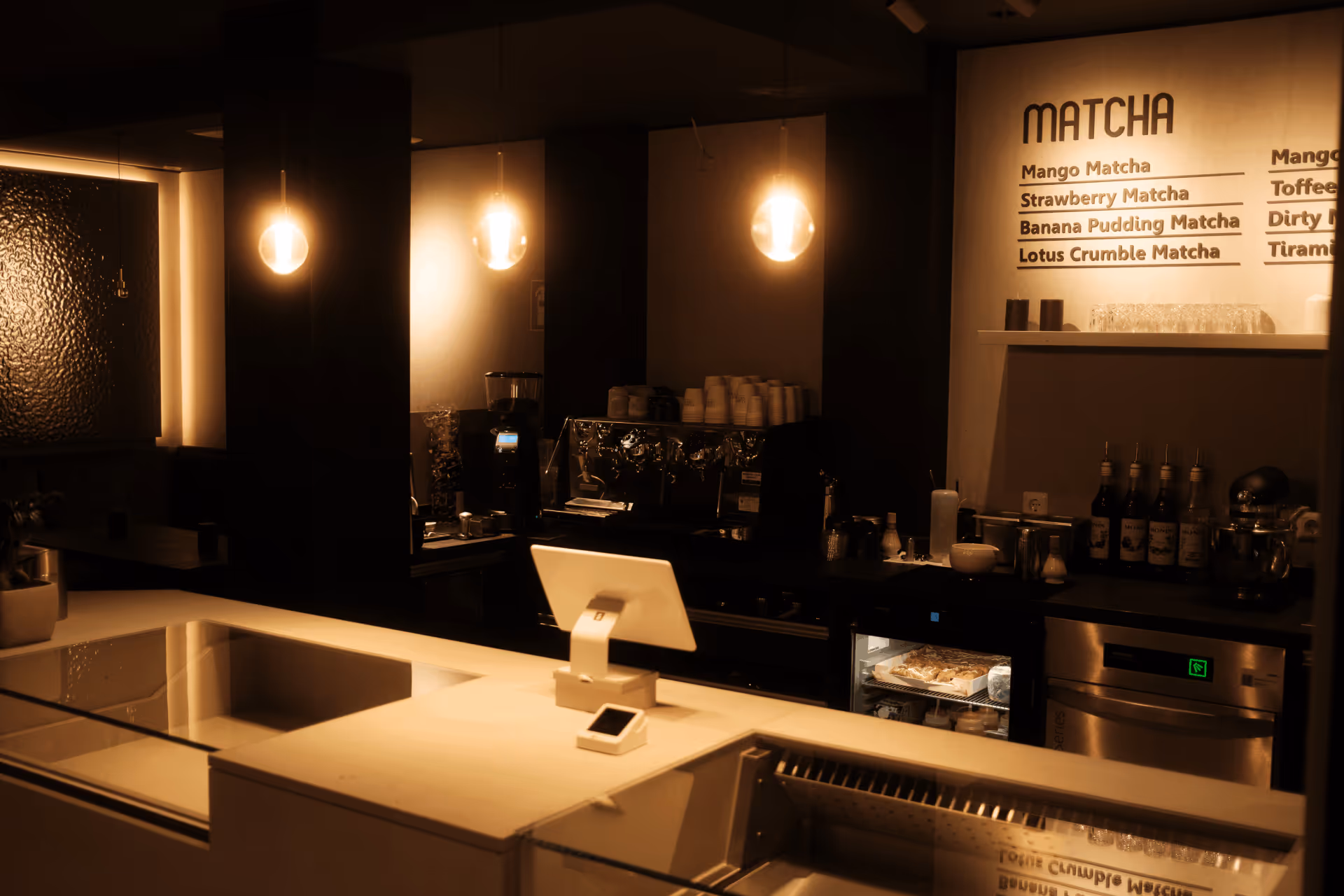 Dunkel beleuchtete moderne Kaffee- und Matcha-Bar mit Espresso-Maschine, Kasse und Menü an der Wand.