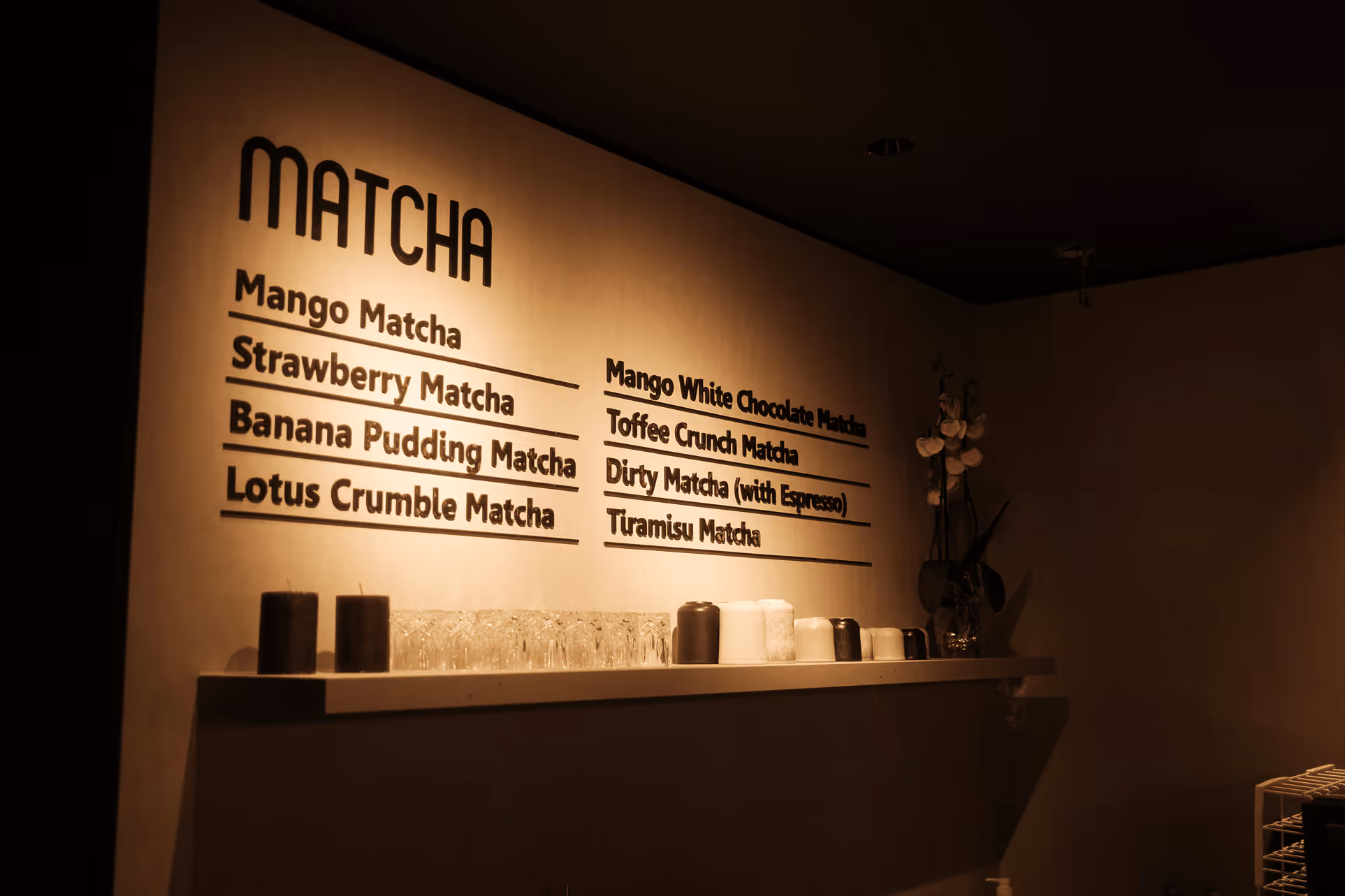 Beleuchtetes Matcha-Menü an einer Wand mit verschiedenen Matcha-Geschmacksrichtungen und Regalen mit Gläsern und Bechern darunter.