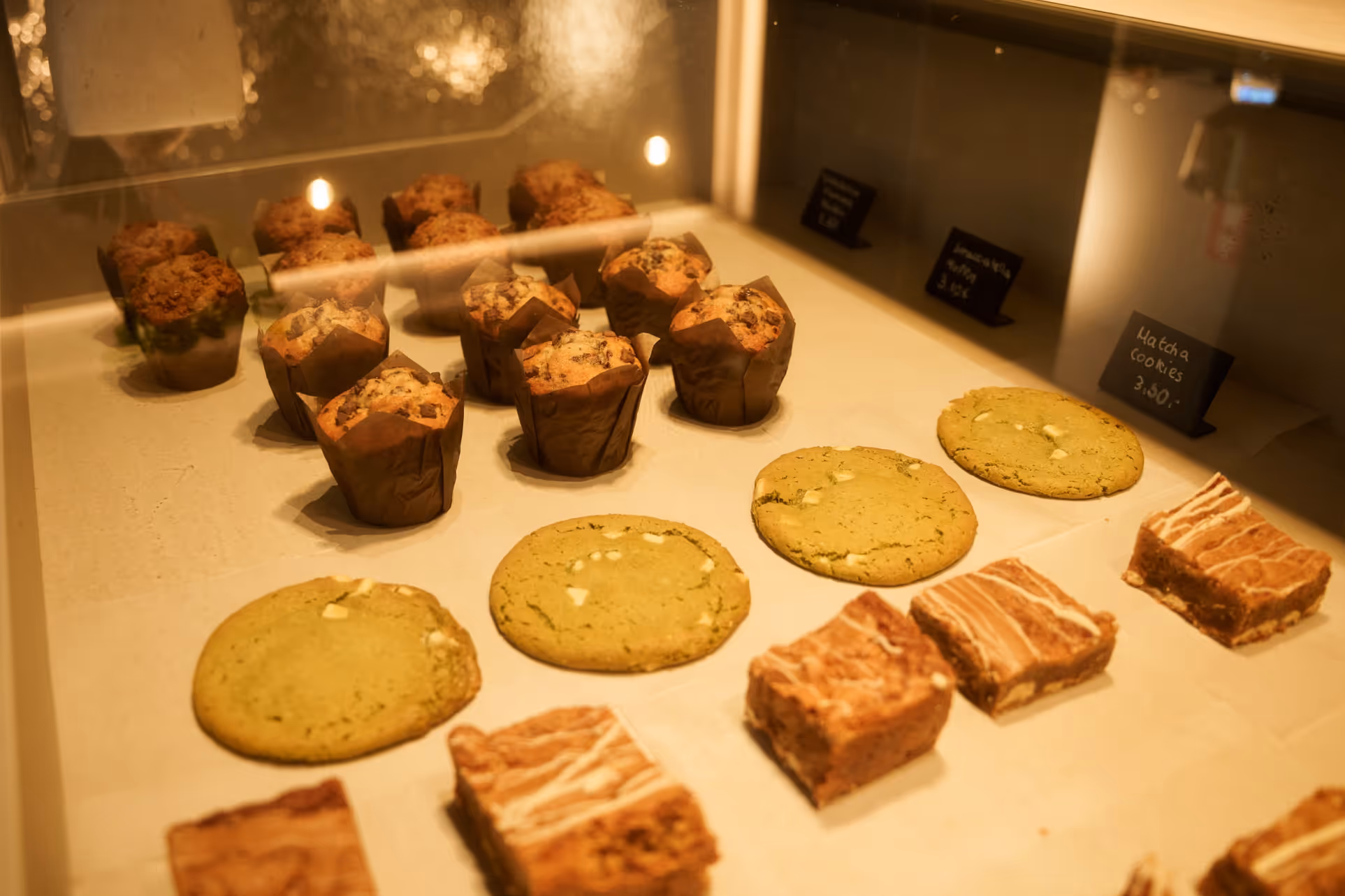 Auslage mit Muffins, grünen Matcha-Keksen und Rechtecken von Gebäck in einer Bäckerei.
