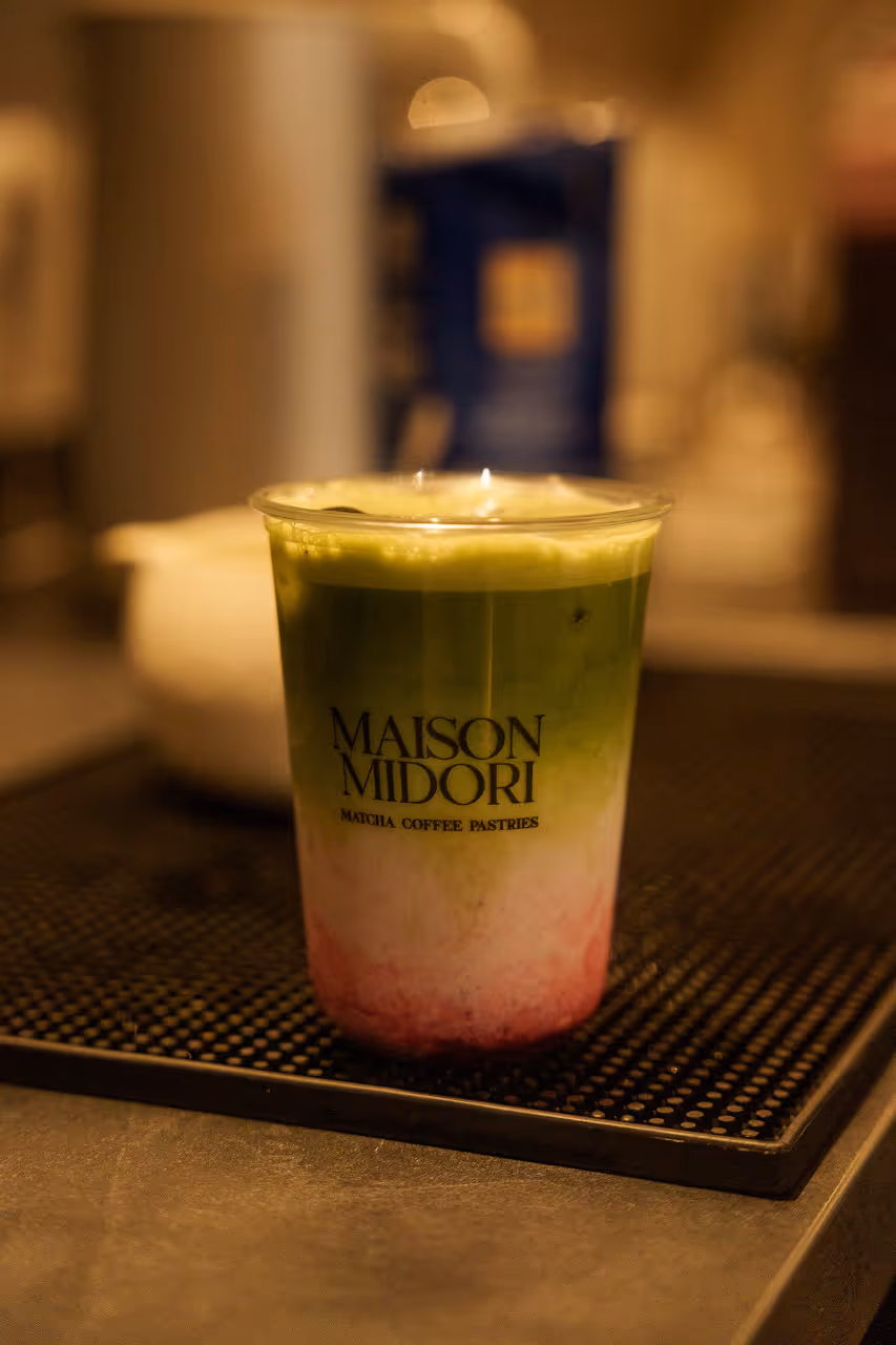 Getränk mit grüner Matcha-Schicht, weißer Mitte und rotem Boden in einem transparenten Becher mit der Aufschrift Maison Midori.