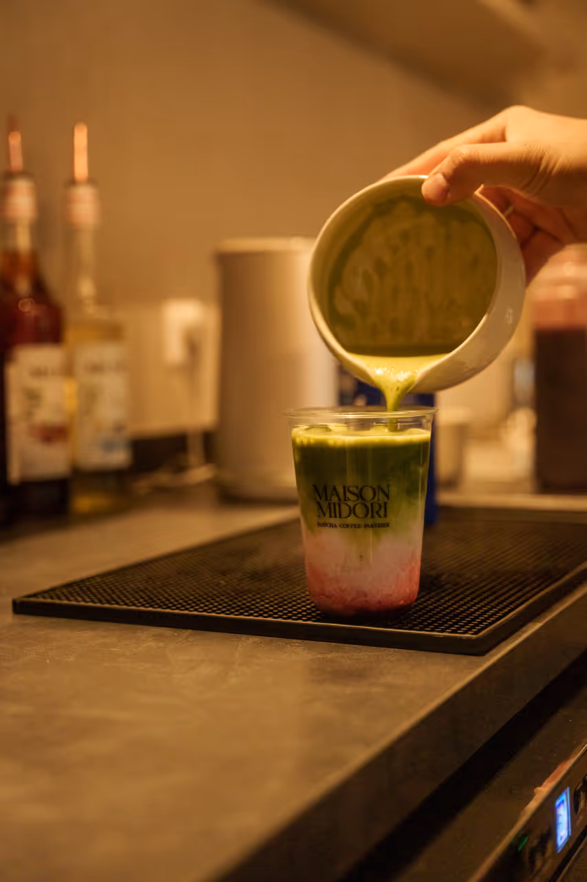 Jemand gießt grünen Matcha-Tee aus einem kleinen Krug in einen transparenten Becher mit rosa und grün geschichtetem Getränk auf einer Theke.