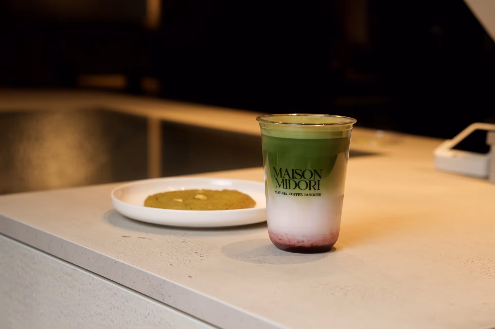 Getränk mit Schichten aus grünem Matcha, weißer Milch und rotem Sirup neben einem Keks auf einem weißen Teller in einem Café.