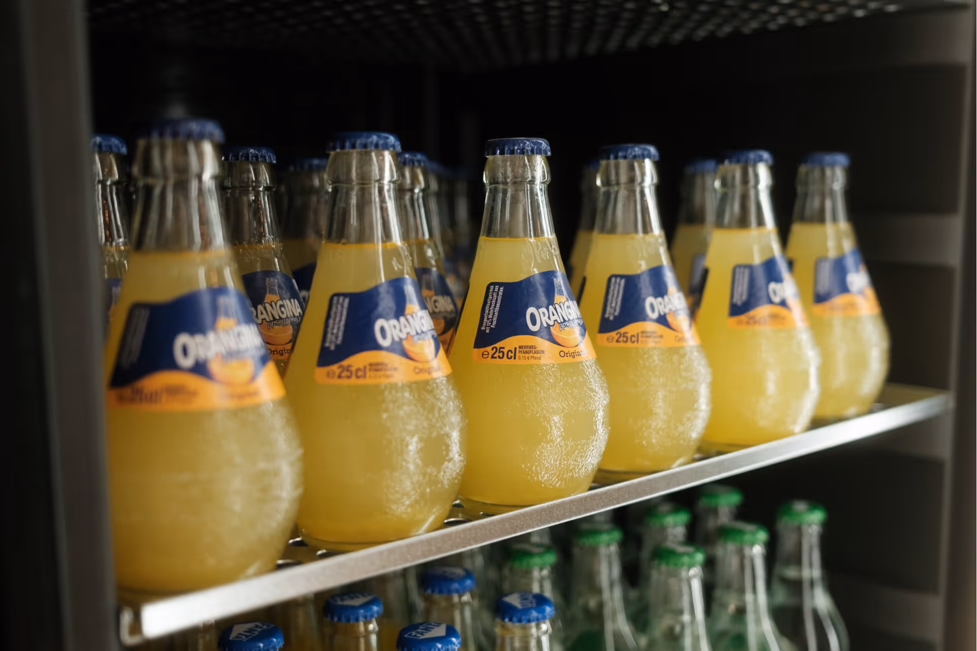 Reihe von Orangina-Flaschen mit gelbem Getränk auf einem Metallregal im Kühlschrank.