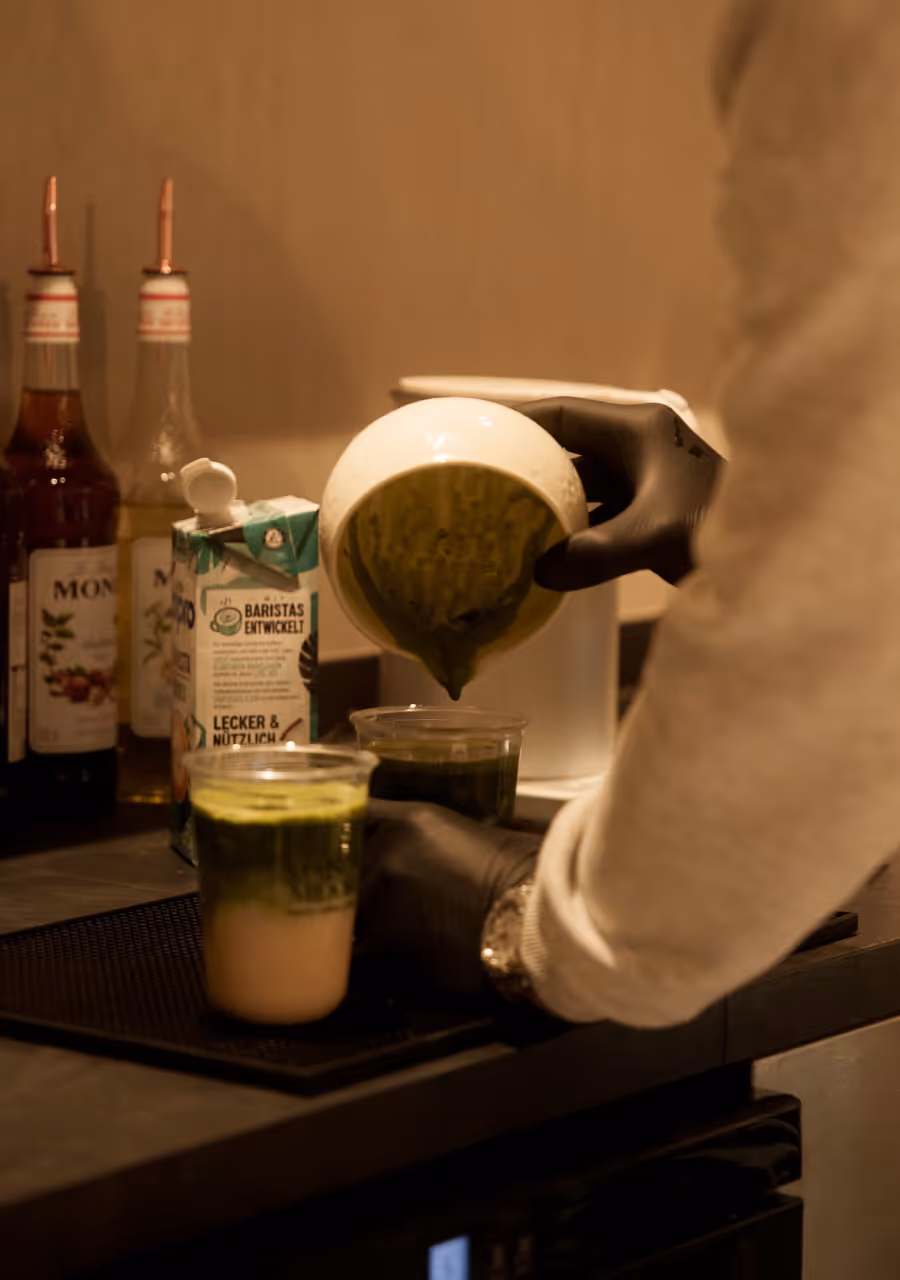 Eine Person mit schwarzen Handschuhen gießt grünes Matcha-Pulver aus einer weißen Schüssel in Plastikbecher mit Milchschaum.