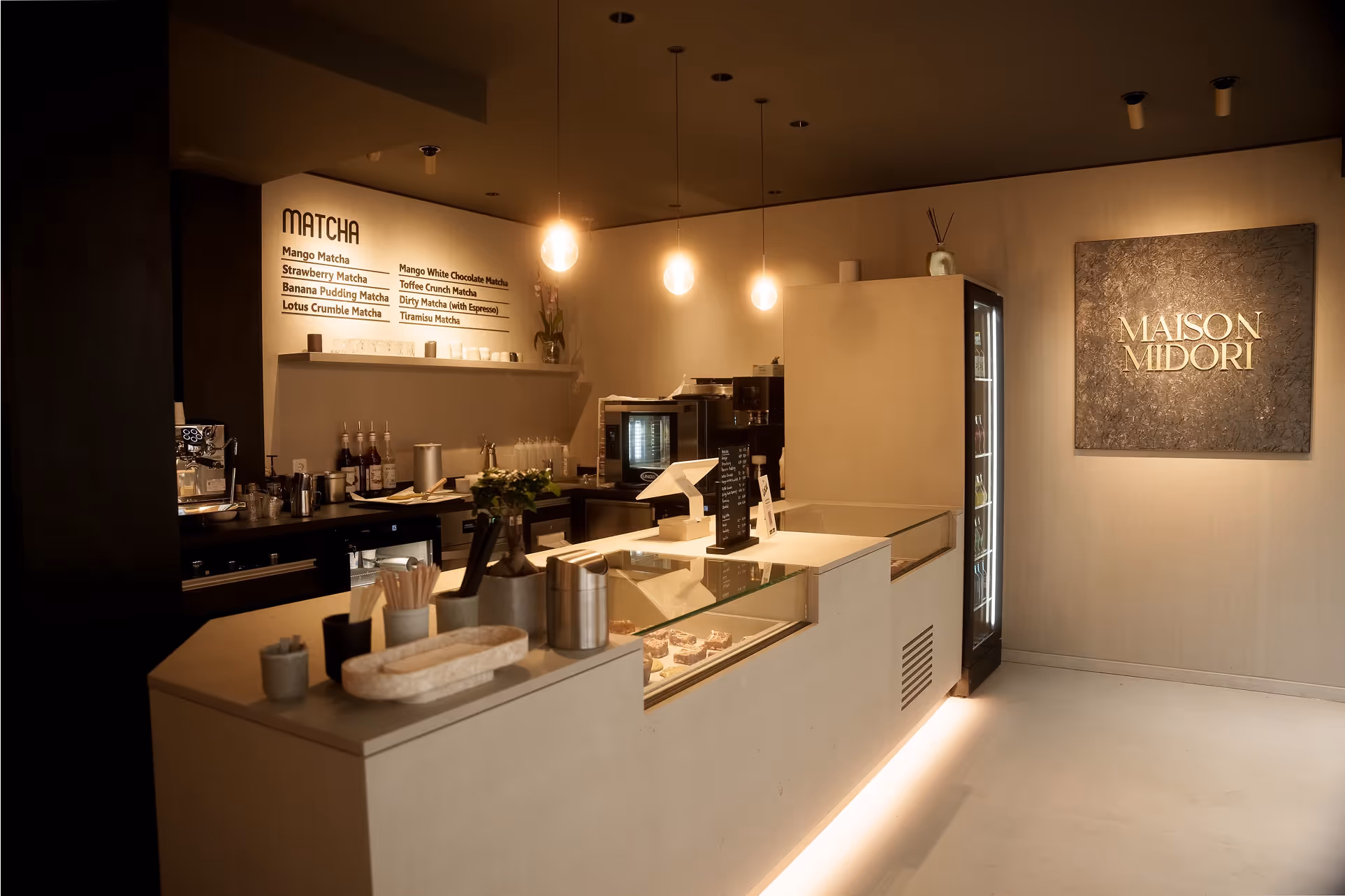 Modernes Caféinterieur mit beleuchtetem Tresen, Menü für Matcha-Getränke und Wandbild mit Aufschrift Maison Midori.