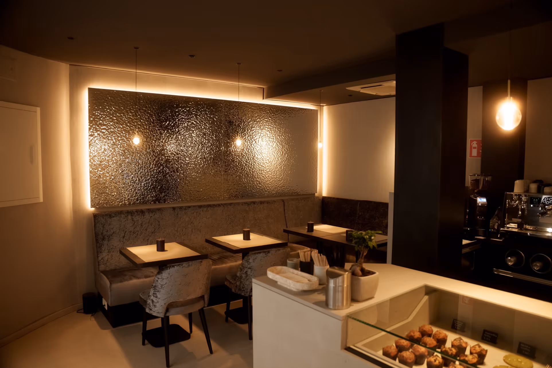 Gemütlicher Caféinnenraum mit gedämpfter Beleuchtung, mehreren Tischen, Stühlen und einer Vitrine mit Gebäck.