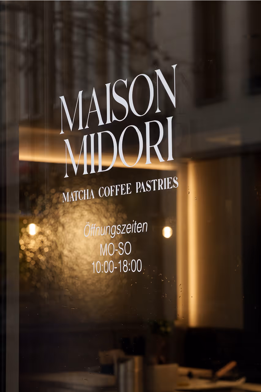 Schaufenster mit Beschriftung von Maison Midori, die Matcha, Kaffee und Gebäck anbietet, sowie Öffnungszeiten Montag bis Sonntag von 10:00 bis 18:00 Uhr.
