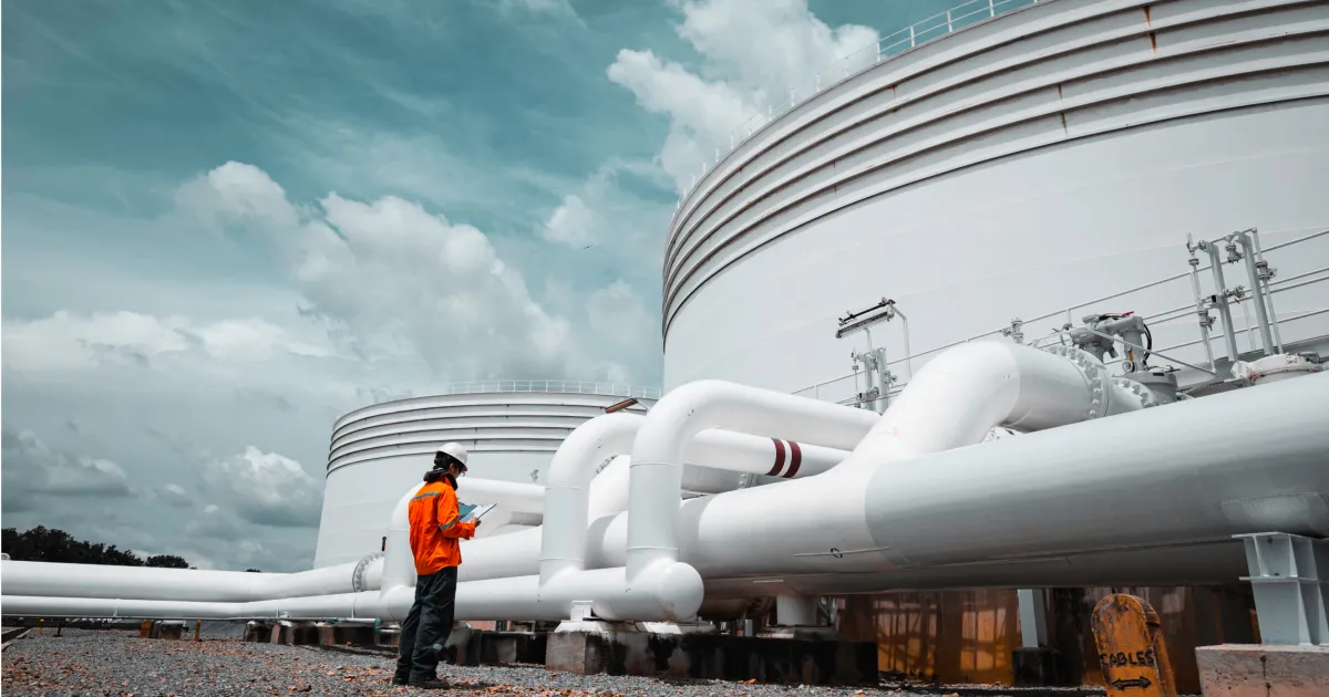 Oil and gas worker in an orange safety suit and hard hat inspecting white storage tanks and pipework at a refinery under a cloudy sky.