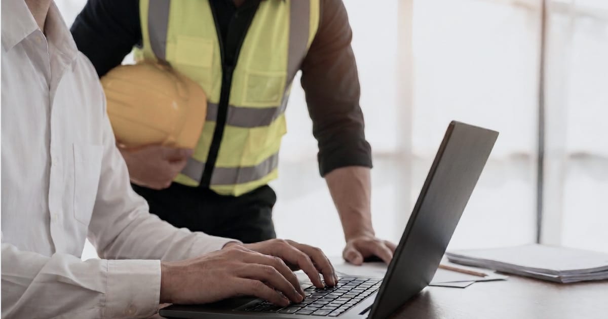 An IT specialist types on a laptop while a maintenance leader in a hi-vis vest holds a hard hat and looks on.
