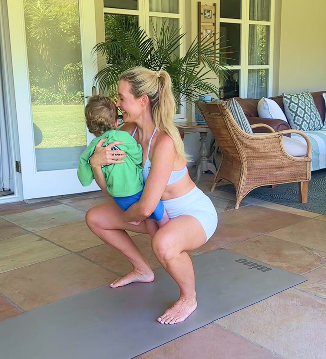Woman in light blue sportswear doing a squat while holding a young child in a green jacket on a yoga mat indoors.