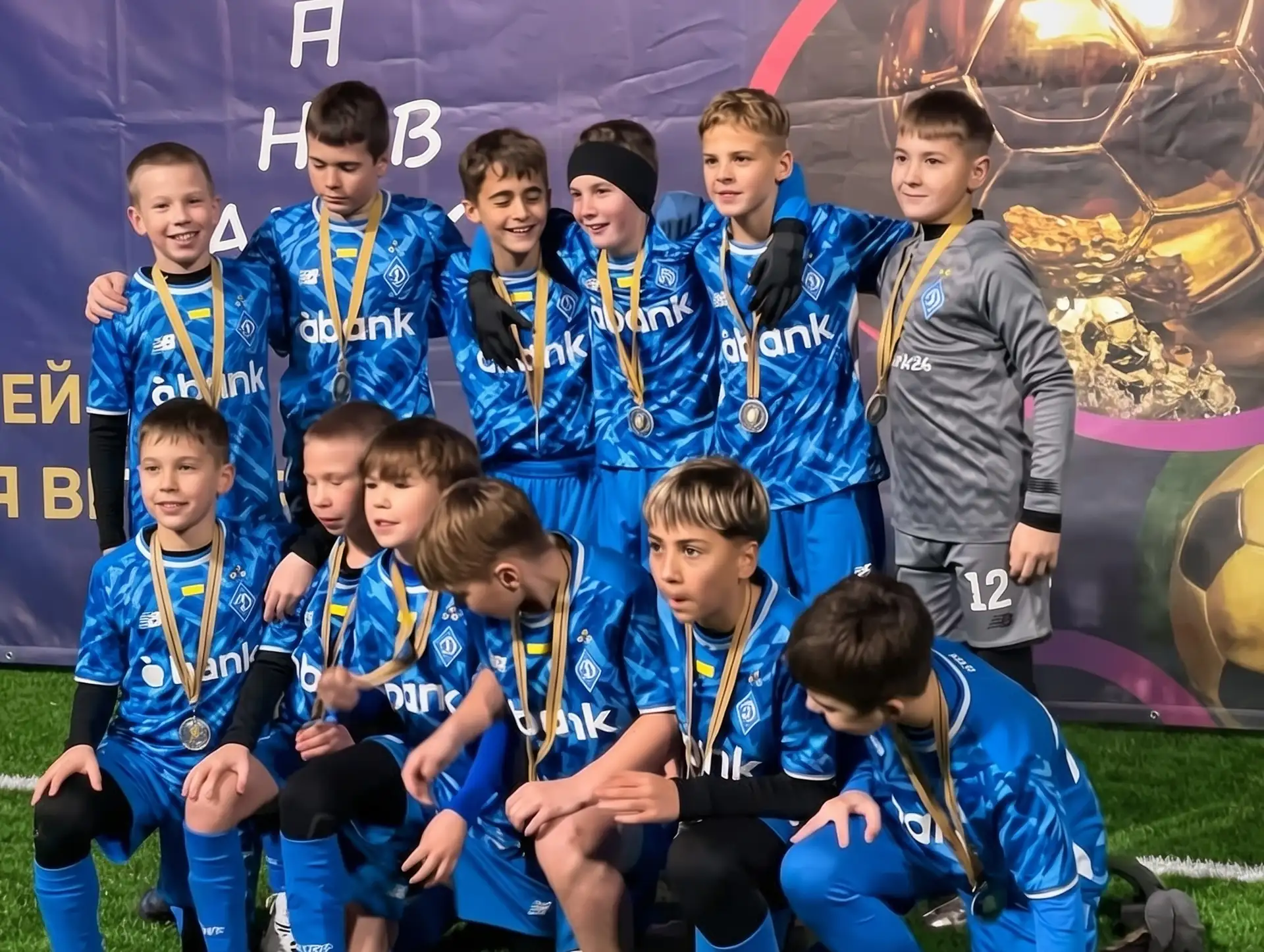 Юні футболісти у синій формі з медалями позують біля банера Кубка володарів золотого мʼяча.