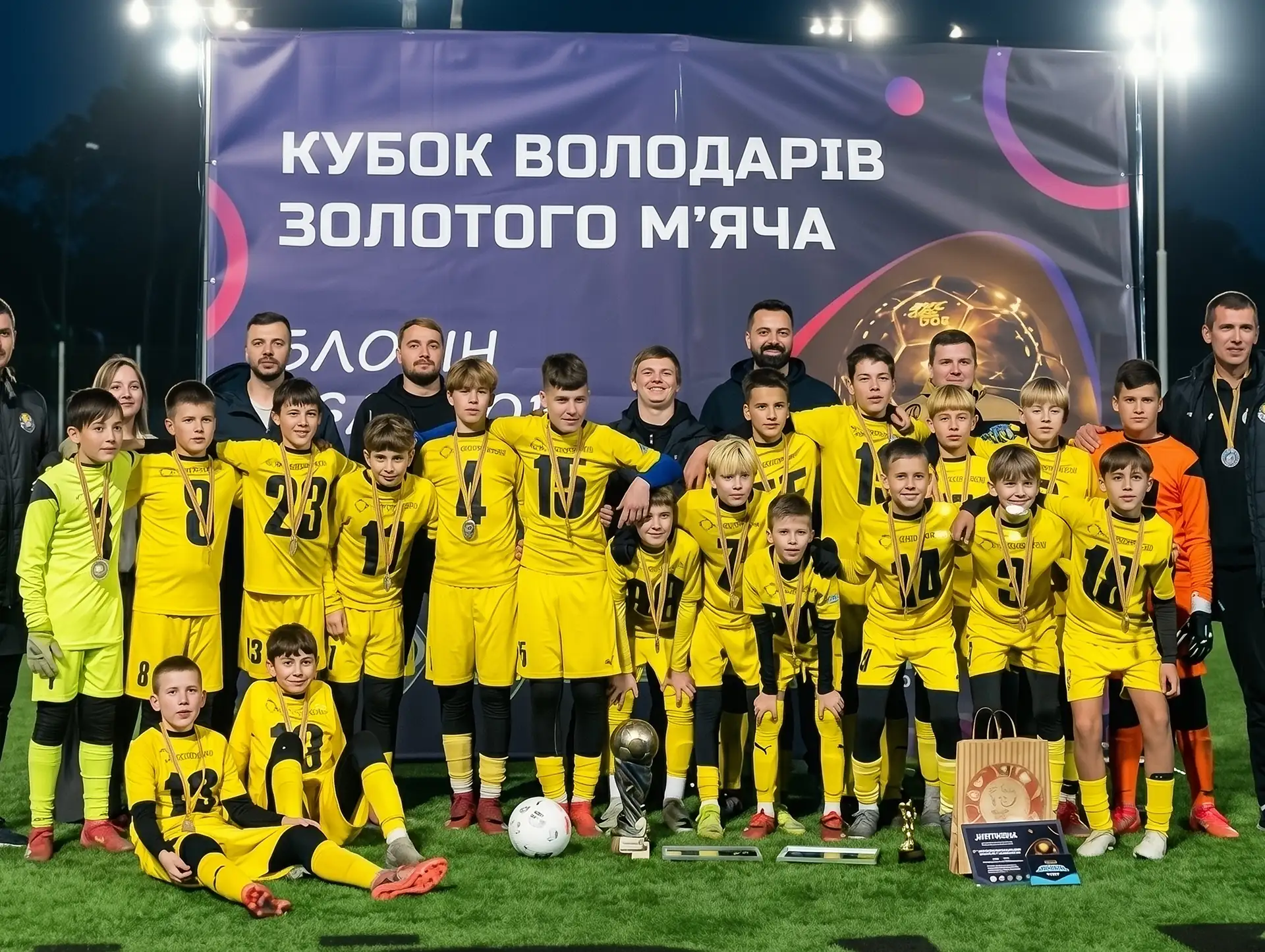 Дитяча футбольна команда в жовтій формі з медалями позує біля банера Кубка володарів золотого мʼяча.