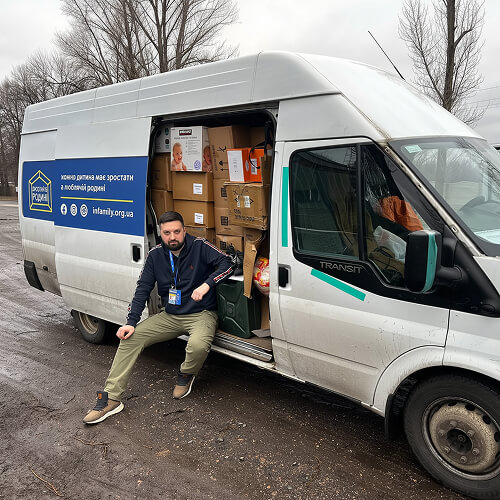 Представник фонду сидить біля автомобіля з коробками гуманітарної допомоги.