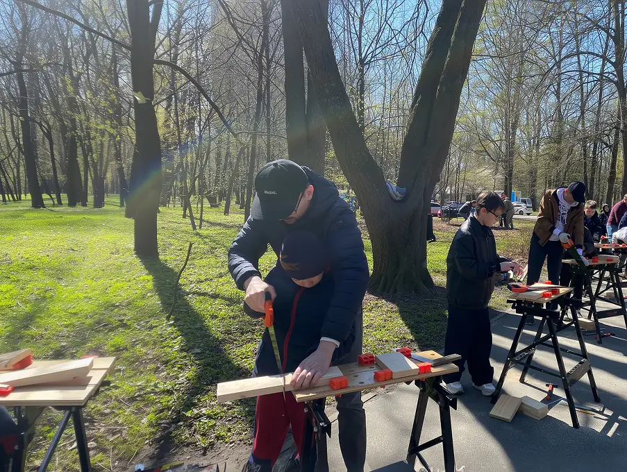 Дорослі та діти майструють деревʼяні вироби на відкритому заході в парку.