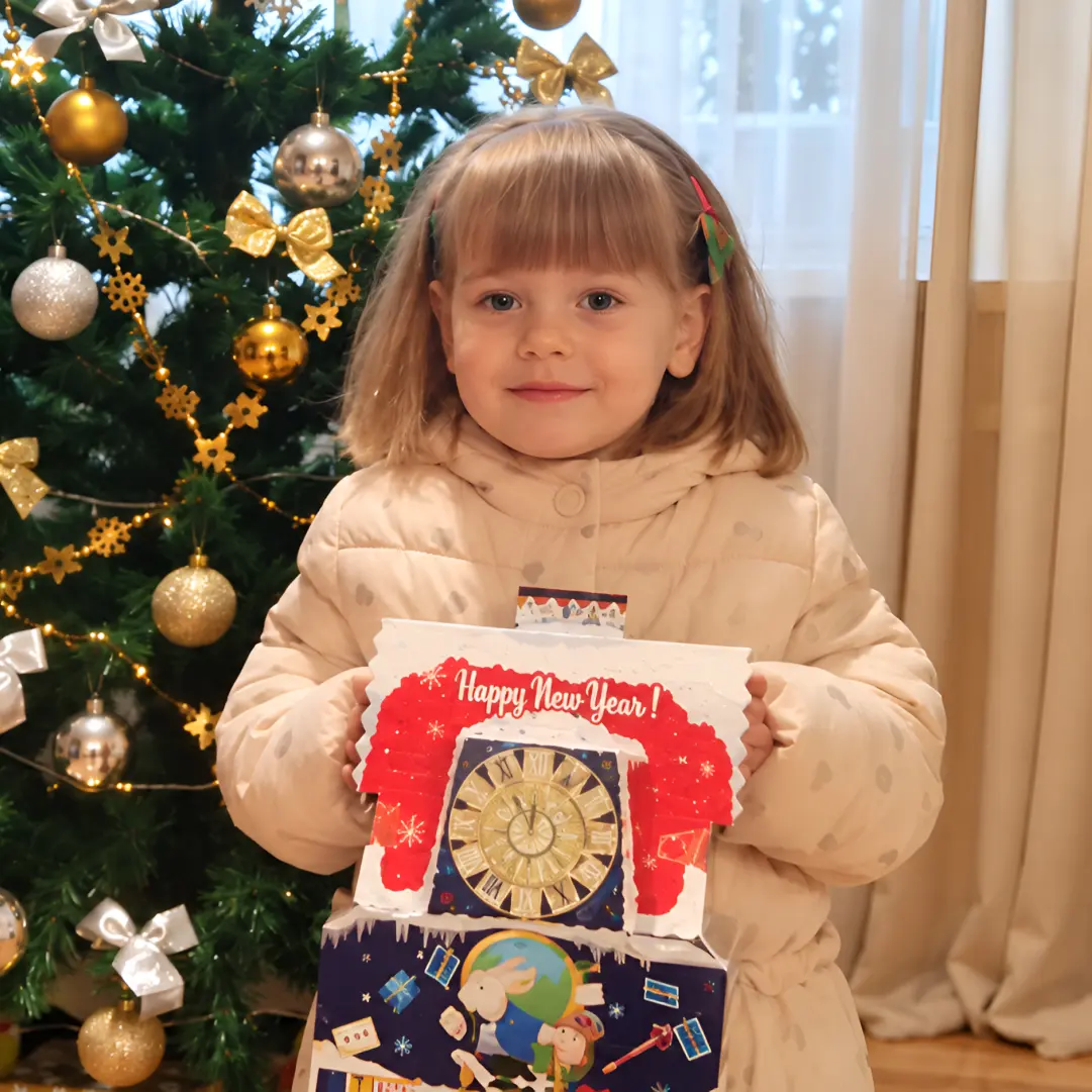 Дівчинка стоїть вдома біля новорічної ялинки з подарунком від команди фонду «Зростай в родині»