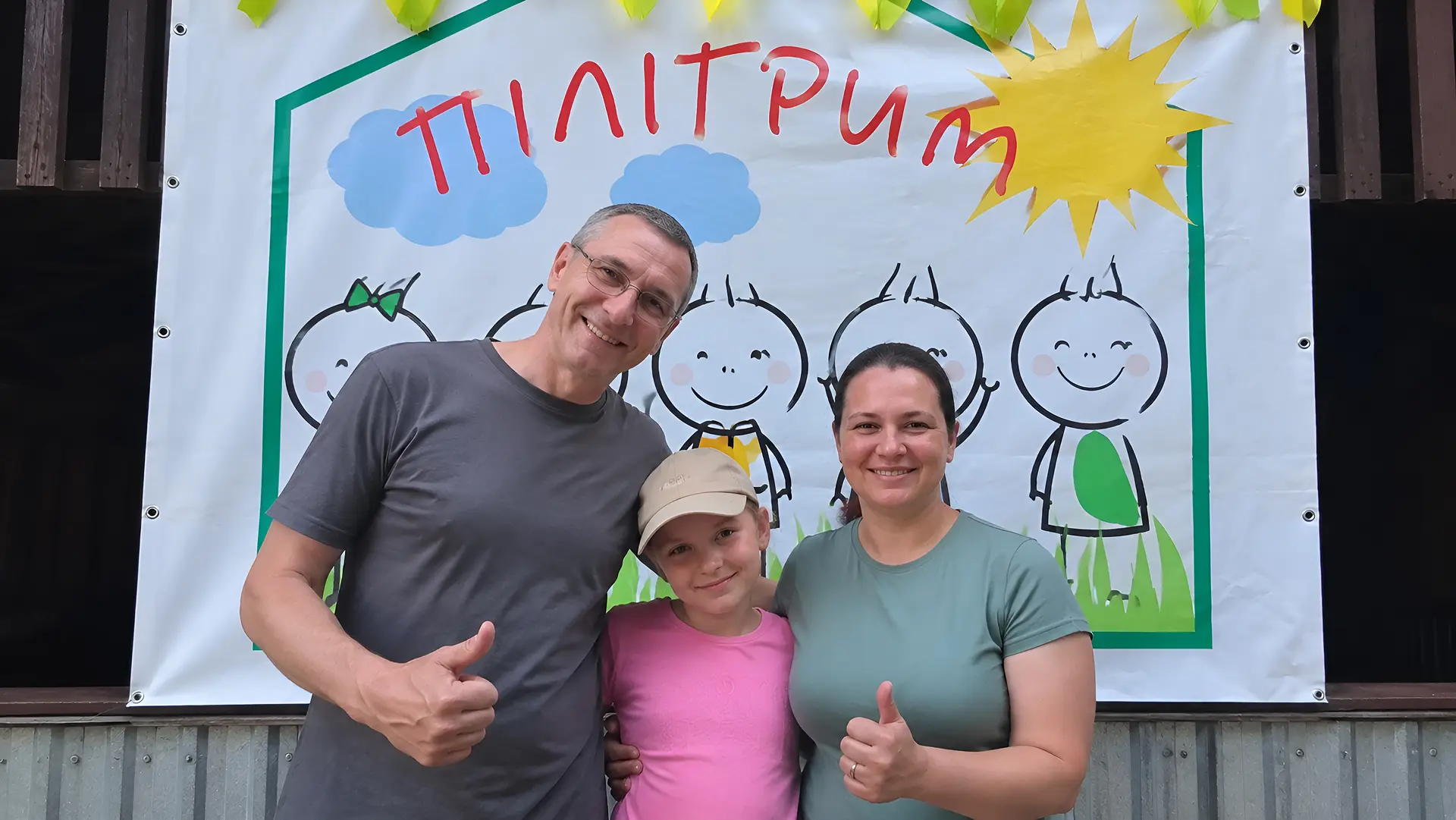 A man, a girl, and a woman in front of a Pilgrim Camp banner.