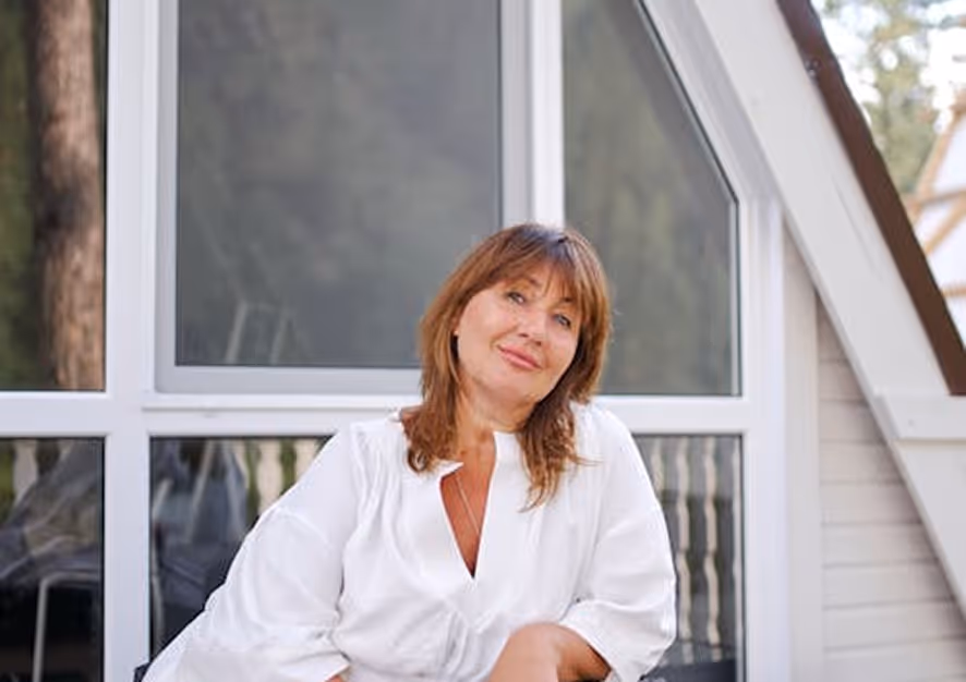 Woman with brown hair wearing a white blouse sitting in front of a large window on a porch.